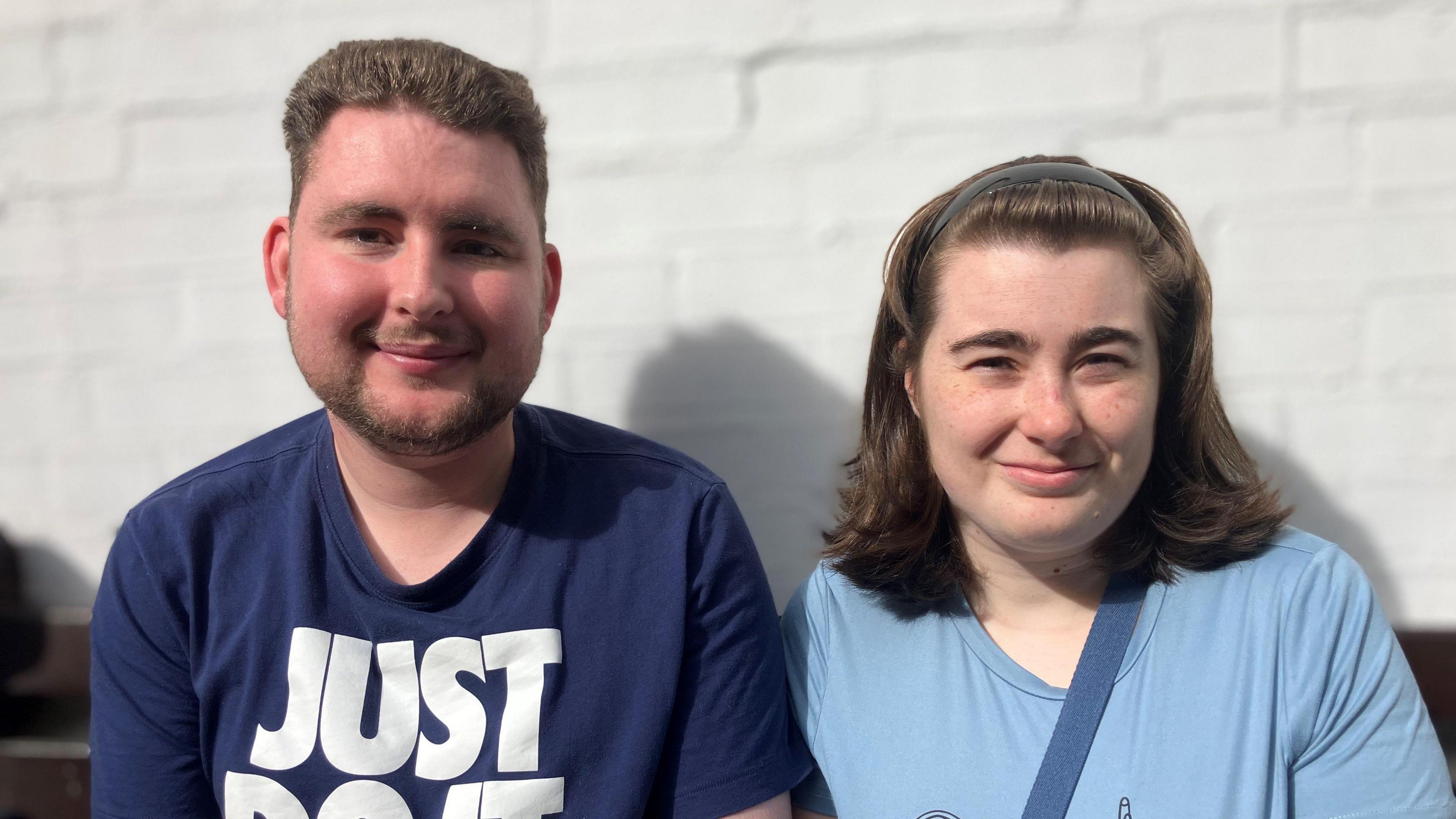 Daniel Strong and Amy Newton. are standing against a white painted wall. They both have brown hair and wearing blue T-shirts. Daniel has a beard.