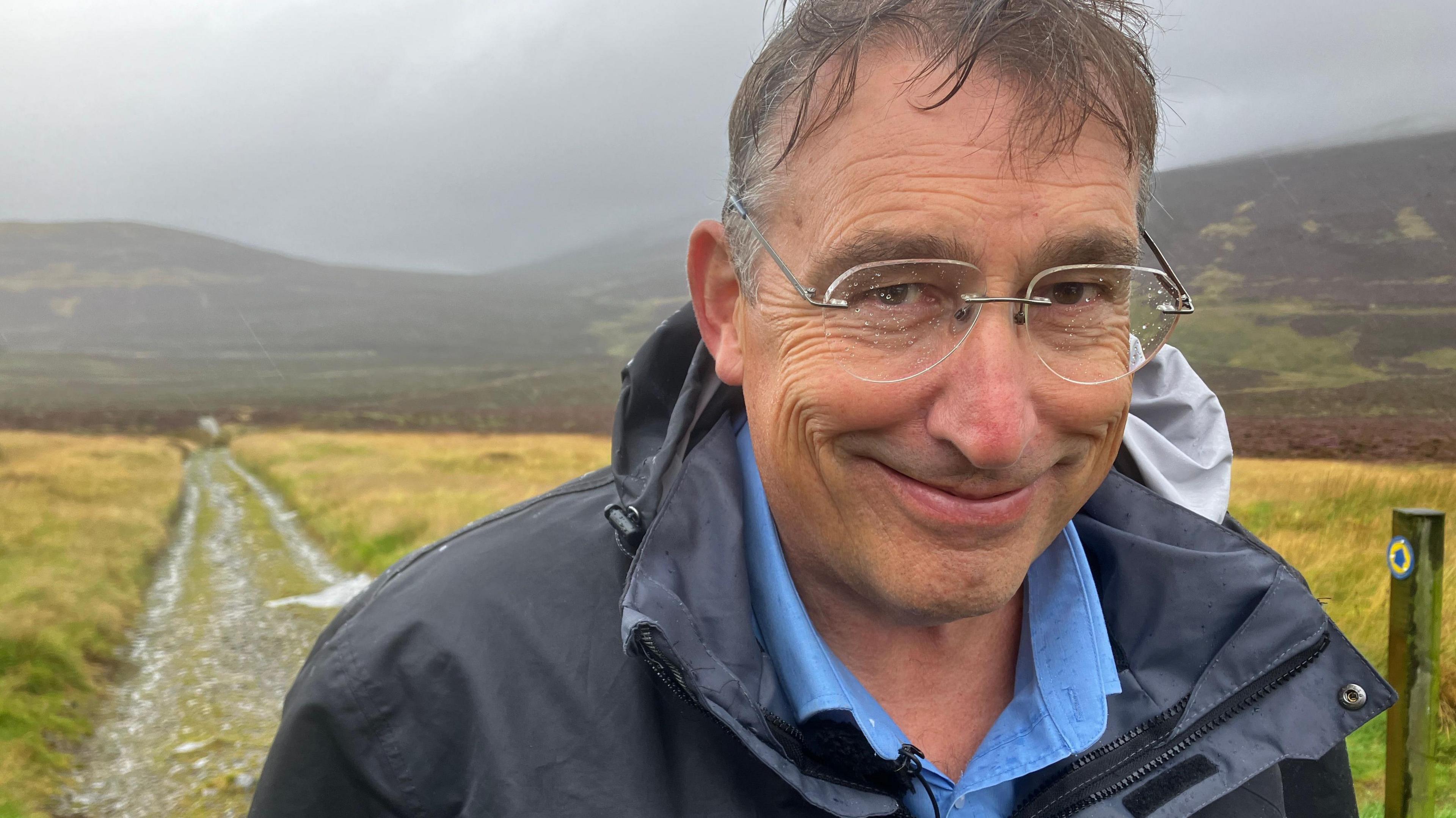 A very wet man with water on his glasses stands in front of Skiddaw 