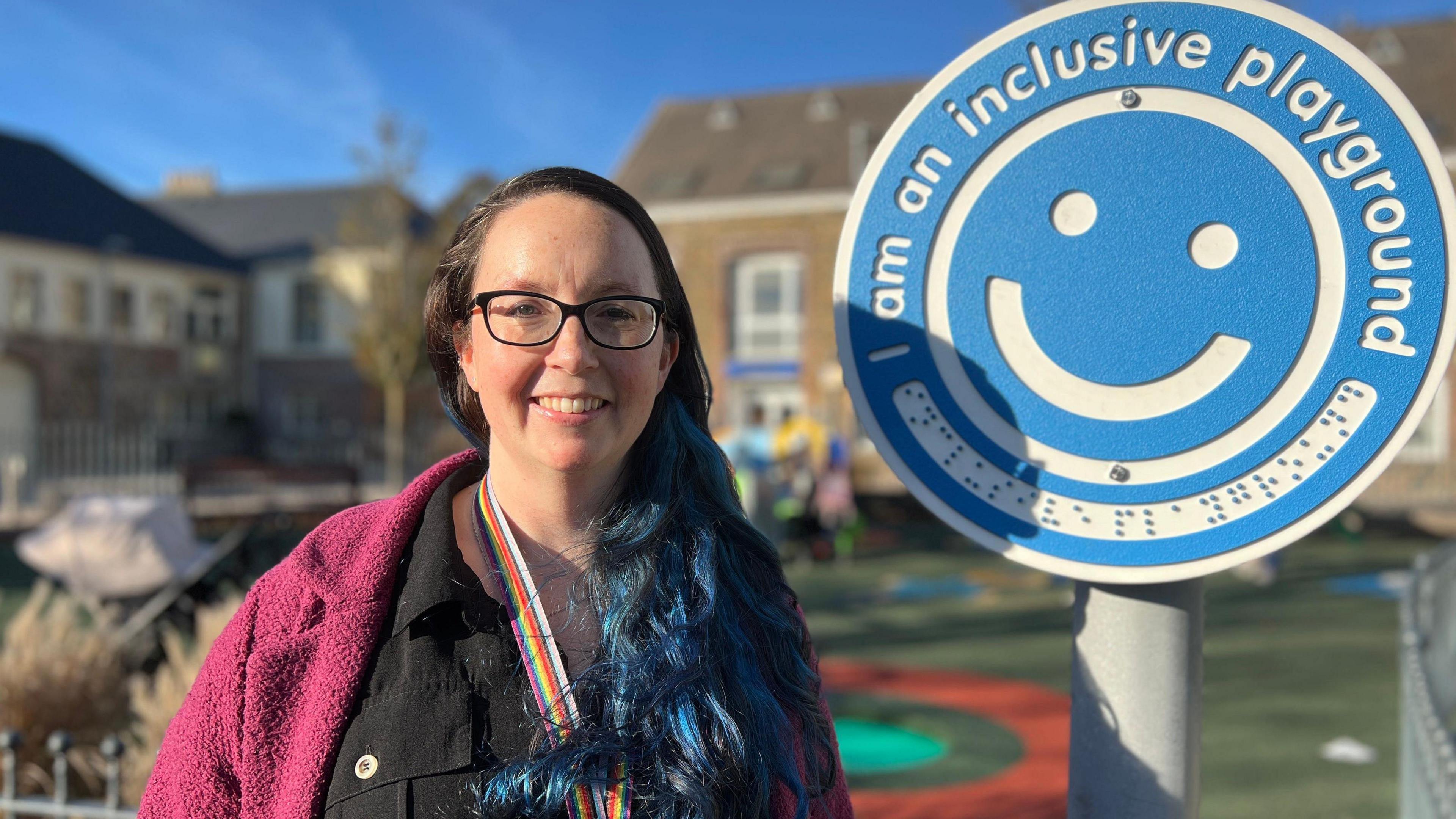 Louise is standing next to a blue circular sign attached to a pole. The sign features a smiling face and the text “I am an inclusive playground.” Louise is wearing a dark outfit with a bright pink outer garment and a rainbow-colored lanyard. Behind her is a fenced playground with colorful play surfaces and buildings in the background.