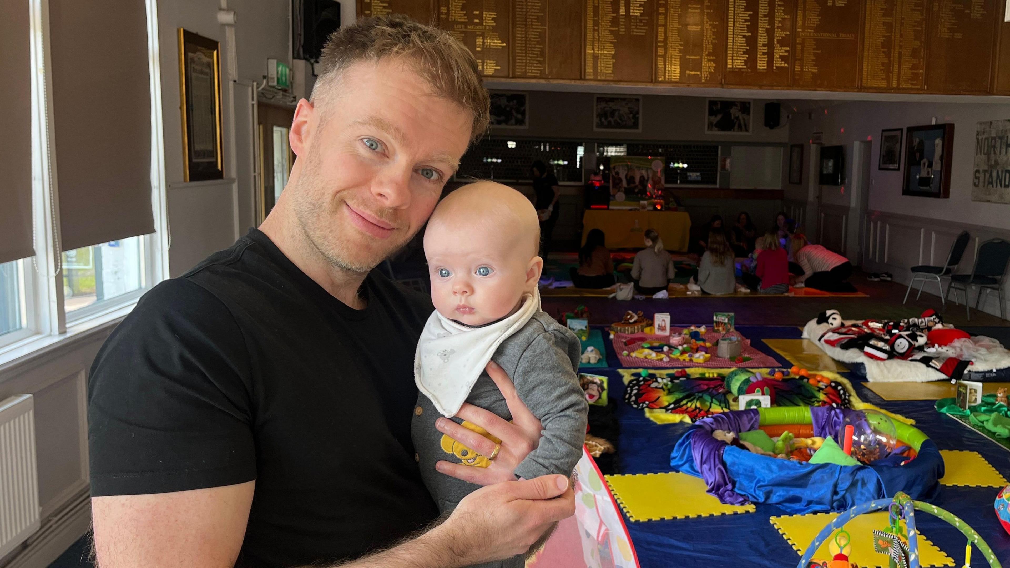 Oli Harrison smiles into the camera holding his baby girl Lily. He is wearing a black T-shirt and has short hair and stubble. Behind them the sensory group class is set out, with lots of colourful mats and toys.