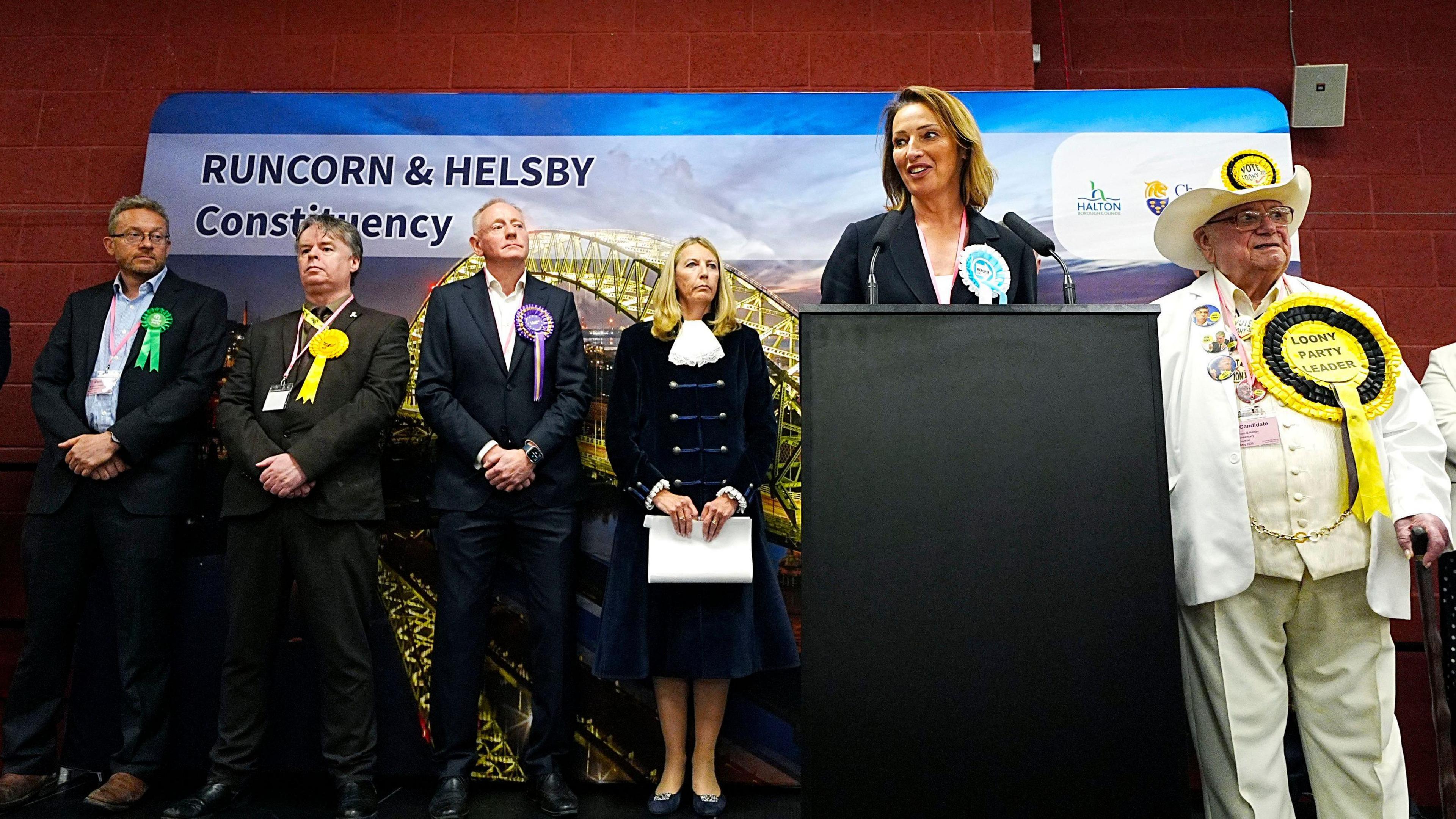 Reform UK MP Sarah Pochin makes her by-election victory speech from a lectern while wearing a white top, dark blue suit and her party's turquoise rosette. Other candidates are in the background along with the returning officer.