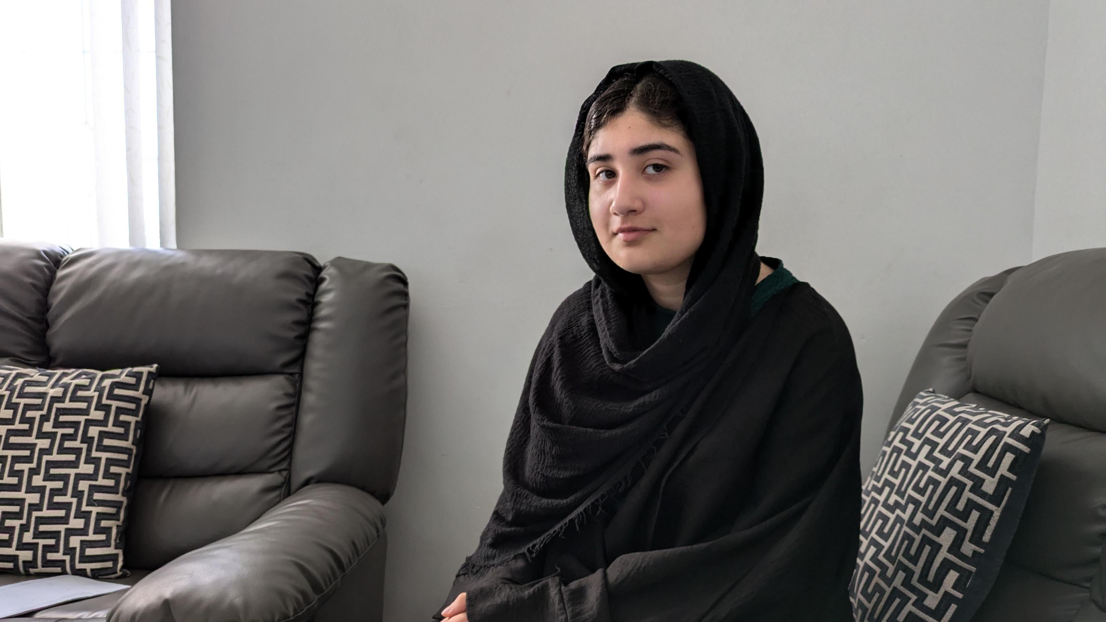 Arooj Hussain, a 19-year-old woman, is sat on a grey leather sofa and is wearing a black headscarf.