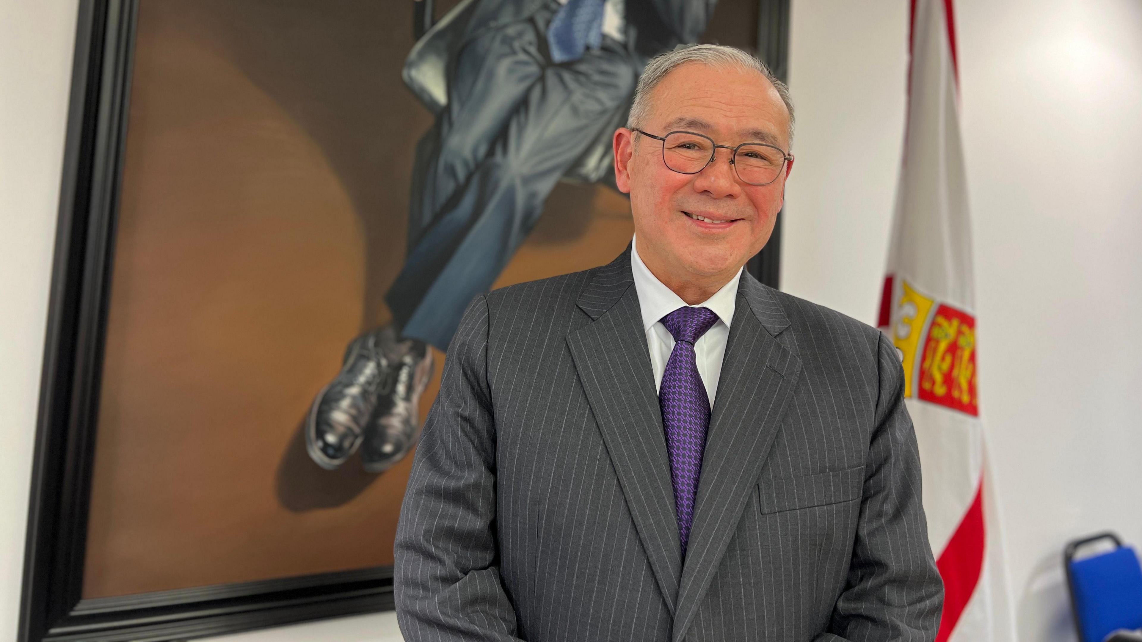 Teodoro smiles at the camera. He's standing in front of an oil painting and a Jersey flag. The oil painting shows a man's legs sitting down at a chair. Teodoro is wearing a black suit with white lines and a purple tie with a white shirt. He's wearing glasses with a thin and rectangular frame.