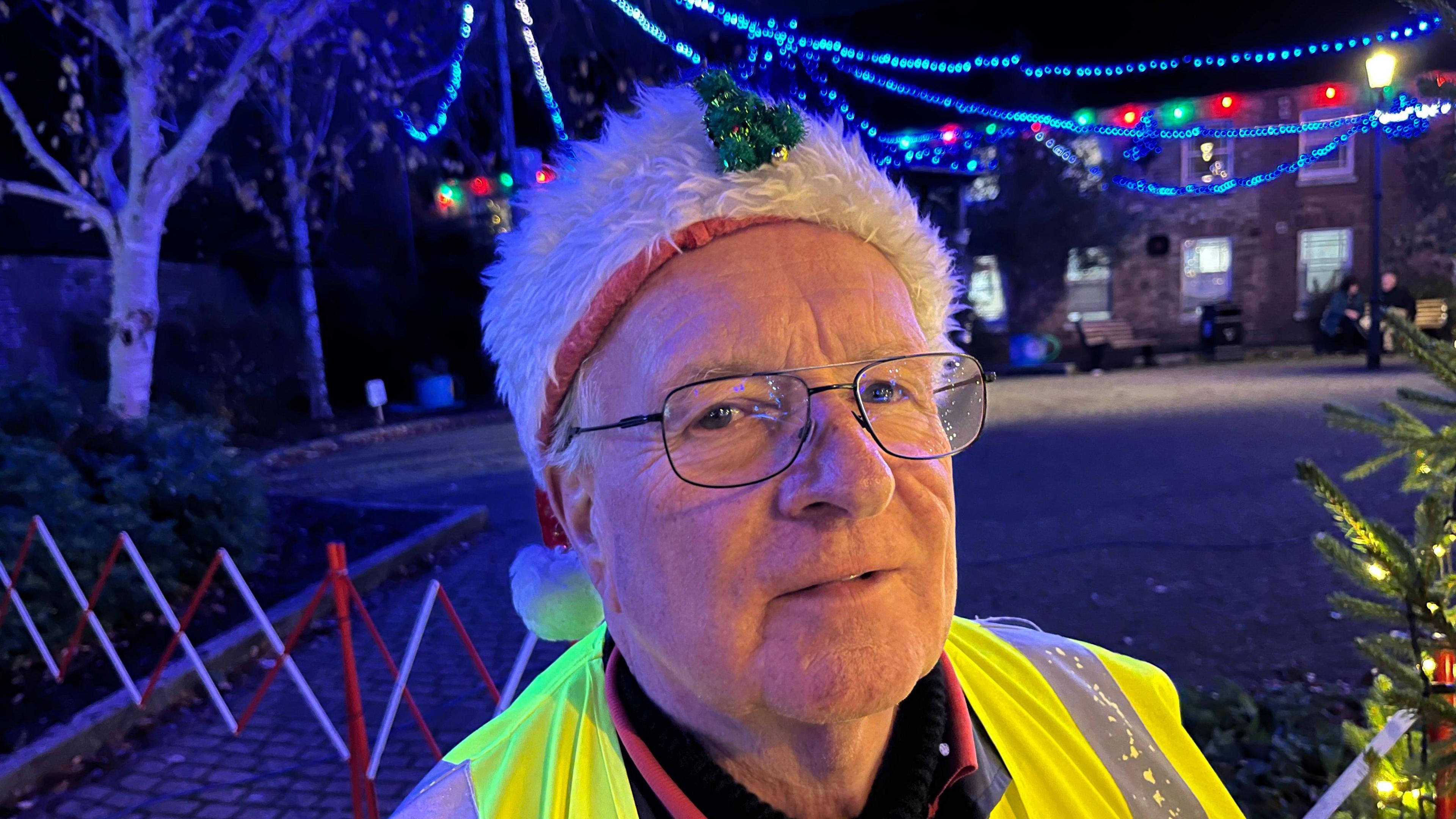 Chris Savill, 72, is wearing a white fur Santa hat with some white hair visible on the side of his head. He wears a high-vis yellow tabard over a dark jumper, gold framed glasses and has a tiny green sparkly Christmas tree on his hat.
Behind him are Christmas lights strung across a path.