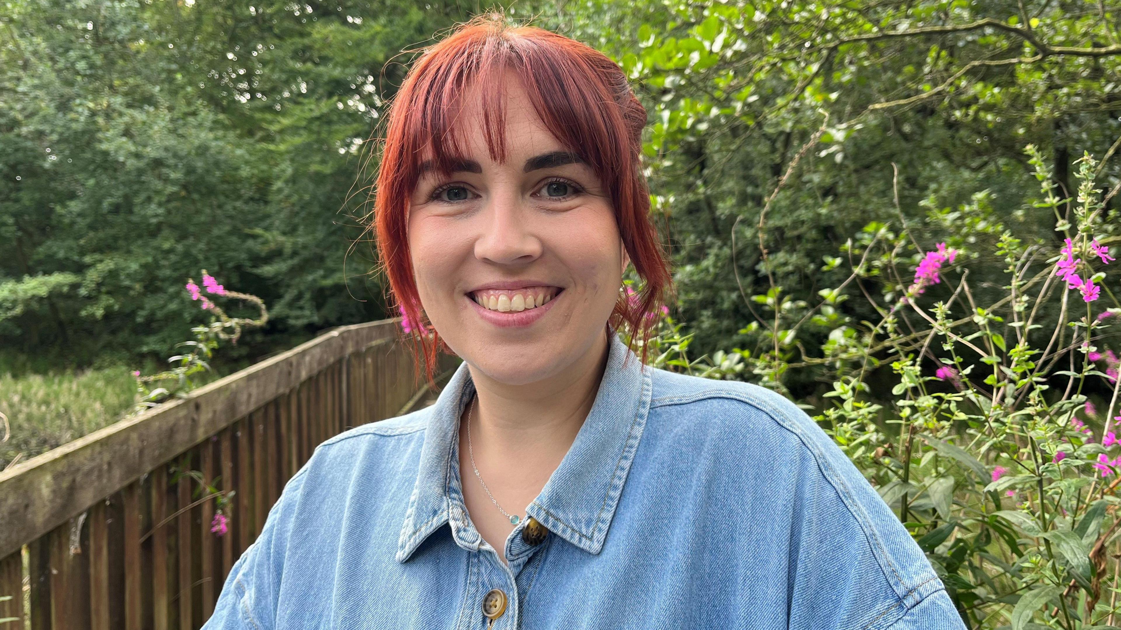 A woman with red hair and a fringe is smiling into the camera, she is wearing a denim jacket, behind her is a wooden bridge.