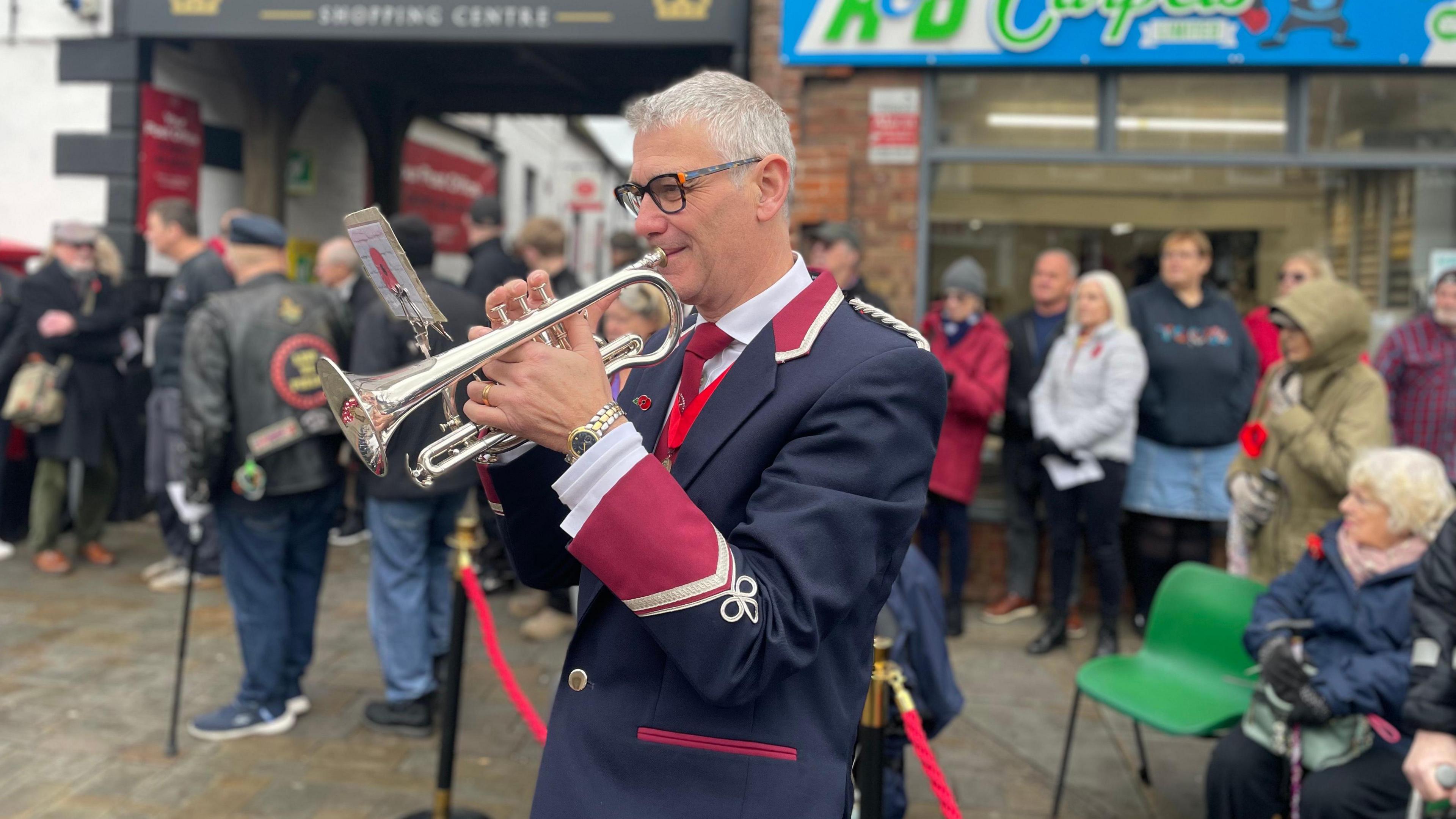 A man is playing the last post on a trumpet wearing a military style blazer which is navy blue with red and gold detail on the sleeves and colour. He is in front of a large crown who are gathered for Armistice Day commemorations in Royal Wootton Bassett. 