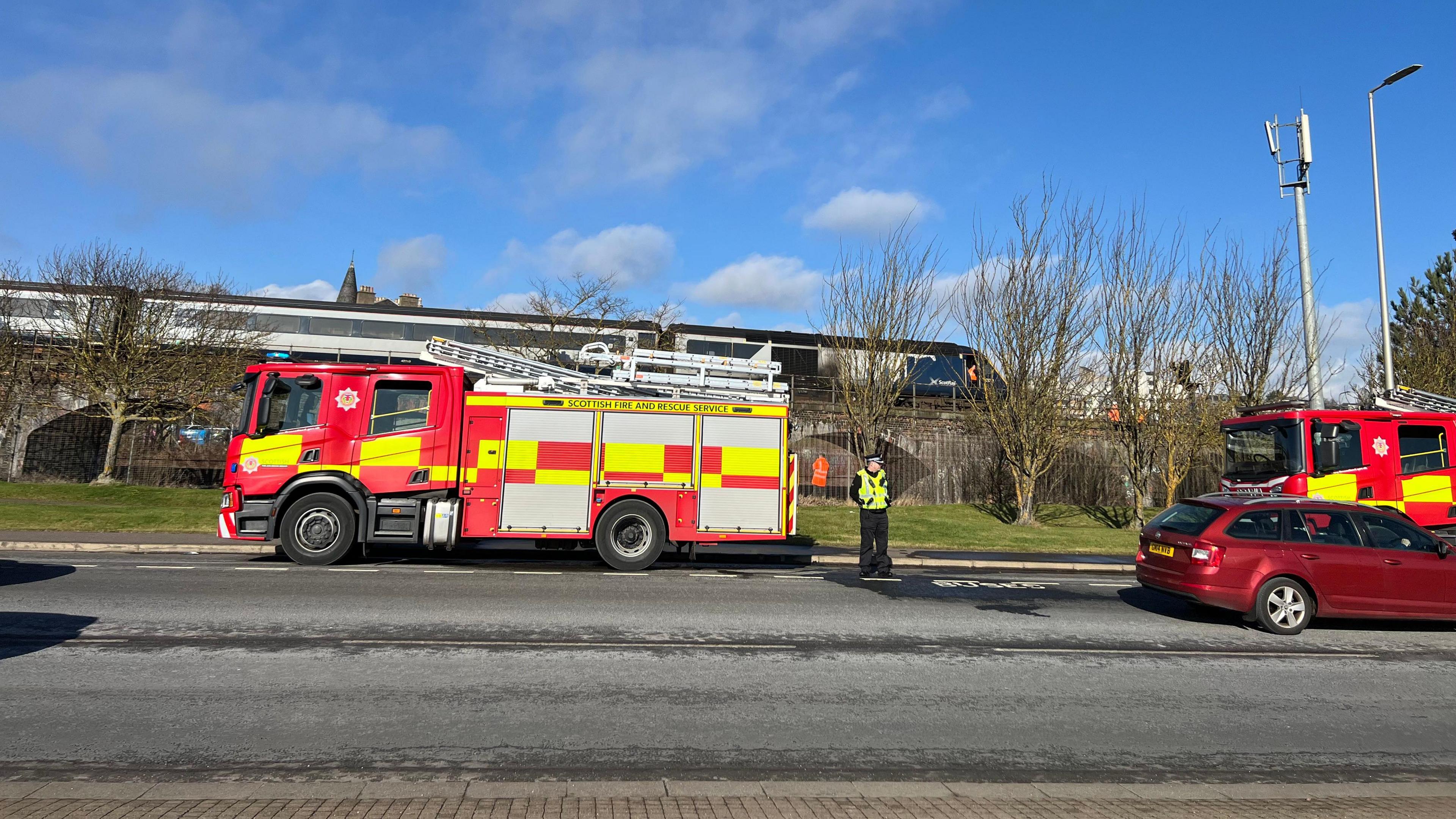 Two red fire engines parked in front of a railway bridge, with a stationary train on the bridge