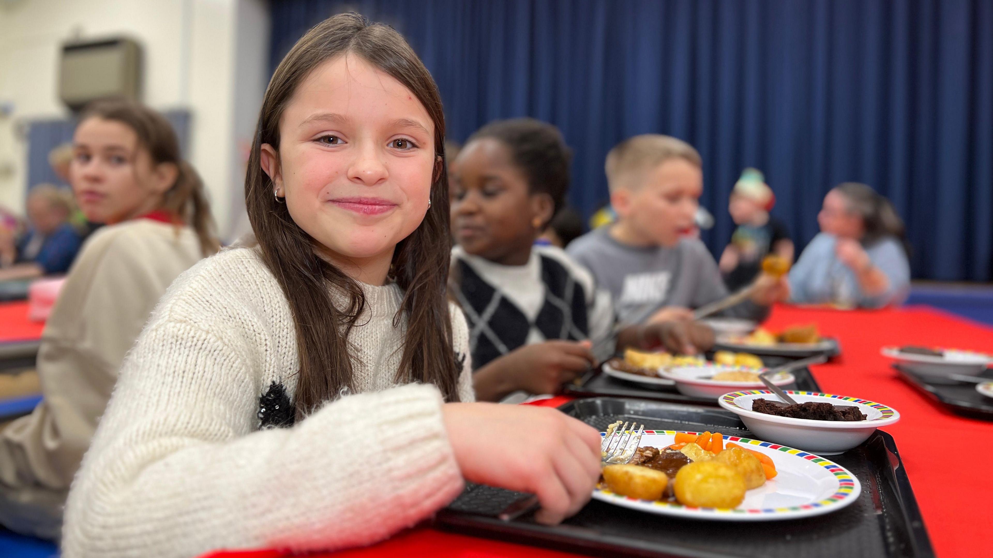 Tilly smiles at the camera as she sits at a children's table with her food on a plate on a tray in front of her. She has dark brown hair which comes over her shoulders and she's wearing a cream coloured knitted jumper.