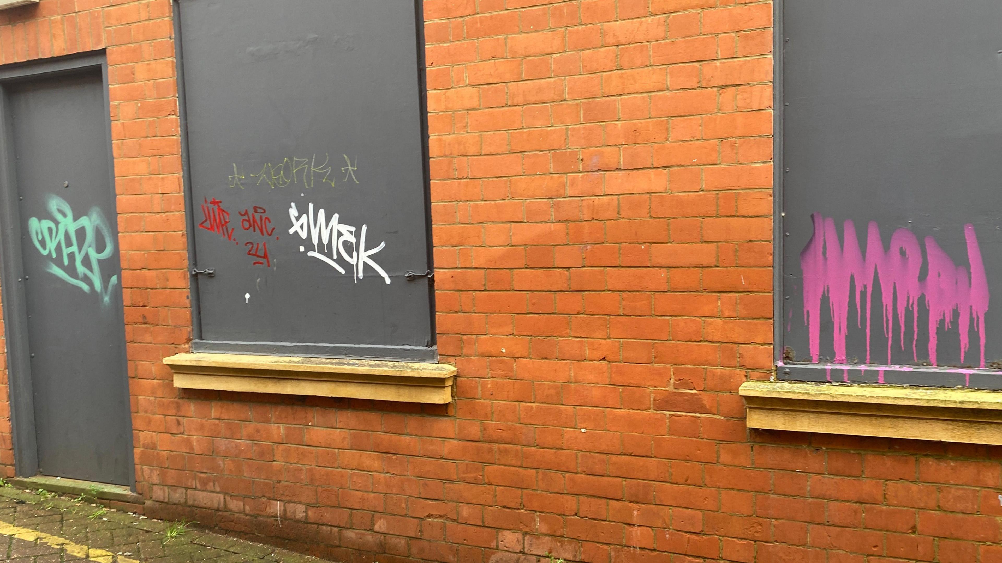 Graffiti has been painted on three grey windows that have been boarded up. The artwork is in lots of different colours.