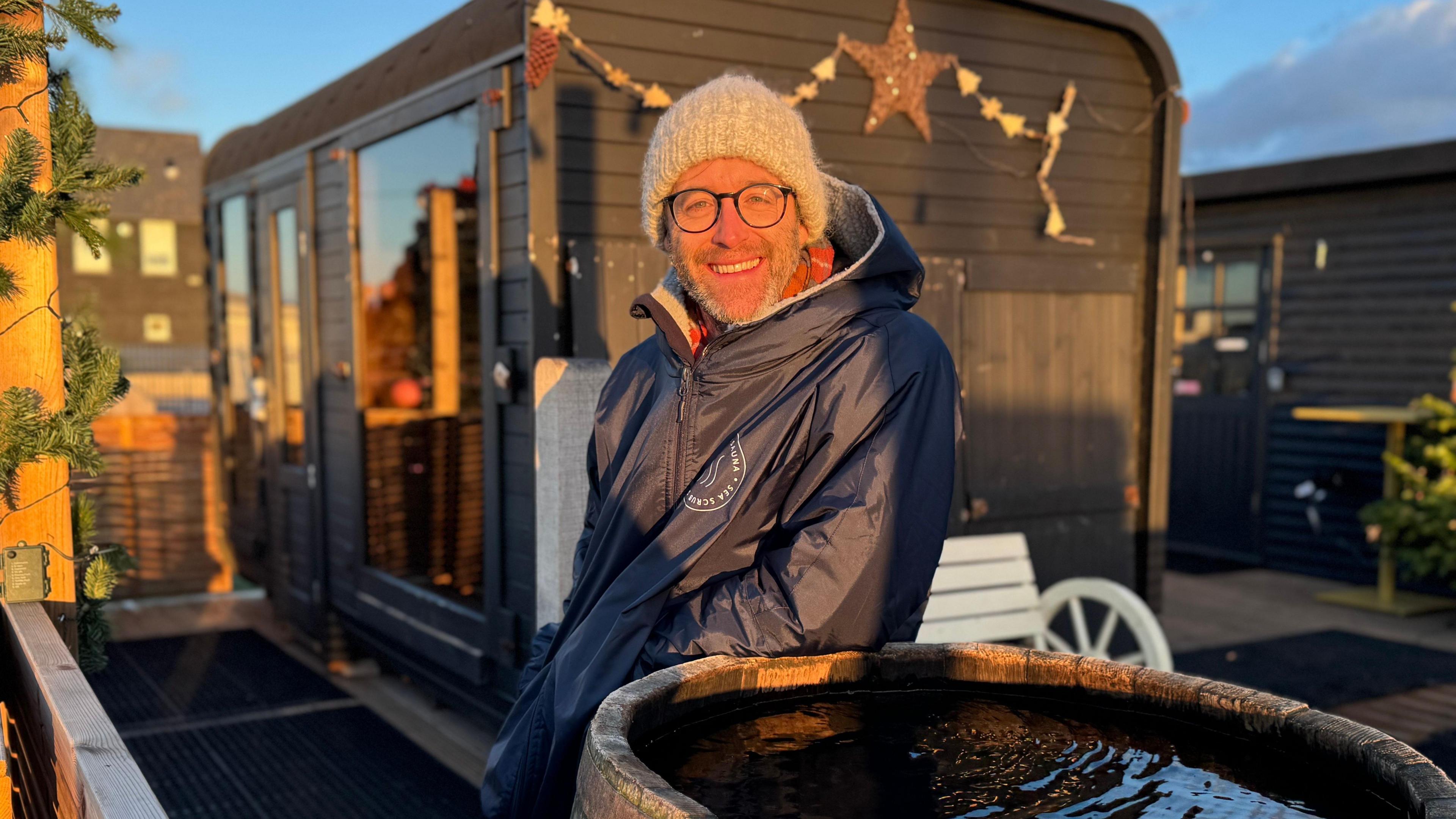 Robin Bartlett wears a grey woolly hat, glasses with black frames and a blue anorak. He stands beside a wooden tub full of water and outside a sauna with black wooden cladding and a glass window.