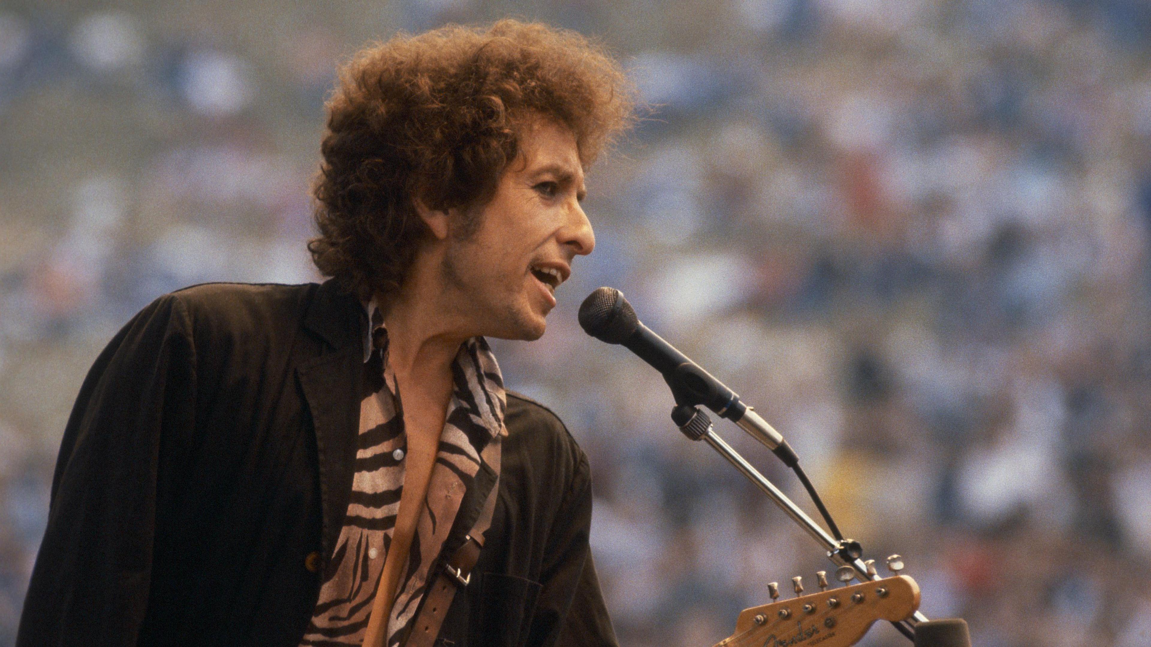 Bob Dylan singing into a microphone, he has curly brown hair and is wearing a black jacket and a tiger print shirt. He is also playing a guitar. 