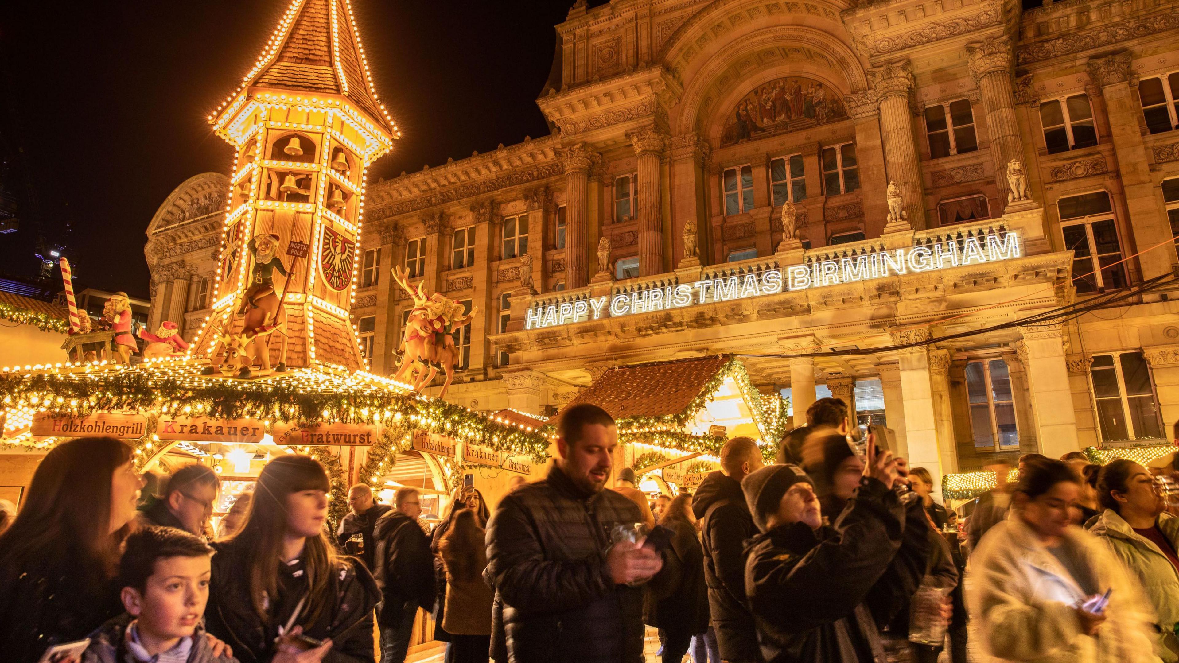 A group of people stood in front of a wooden building and Birmingham City Hall which are lit up with Christmas lights. The lights cover wooden huts while people are dressed in jackets and some hold glasses.