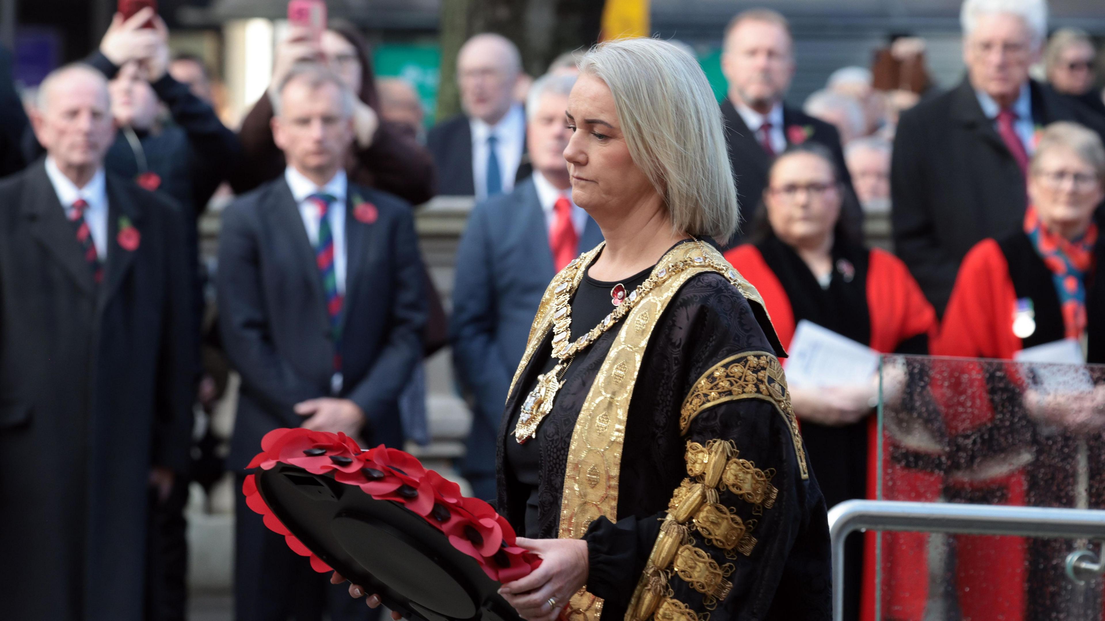 Mayor Tracy Kelly is wearing her official robes. They are black and gold. She's also wearing her gold chain of office and holding a poppy wreath.