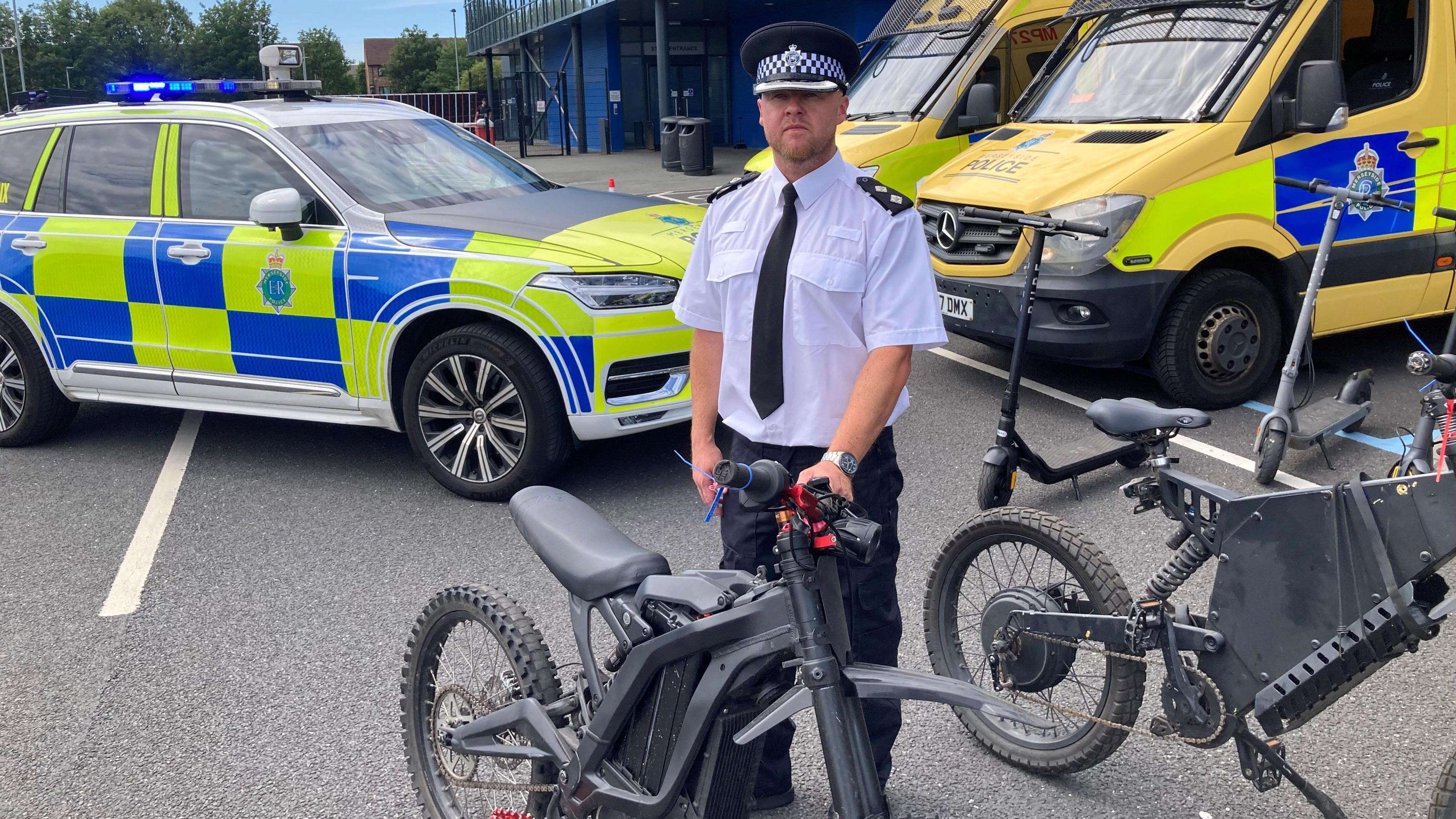 Insp James May, in his police uniform of a black cap, white shirt and black tie, stands behind a black electric bike. There are police patrol cars and yellow police vans in the background.