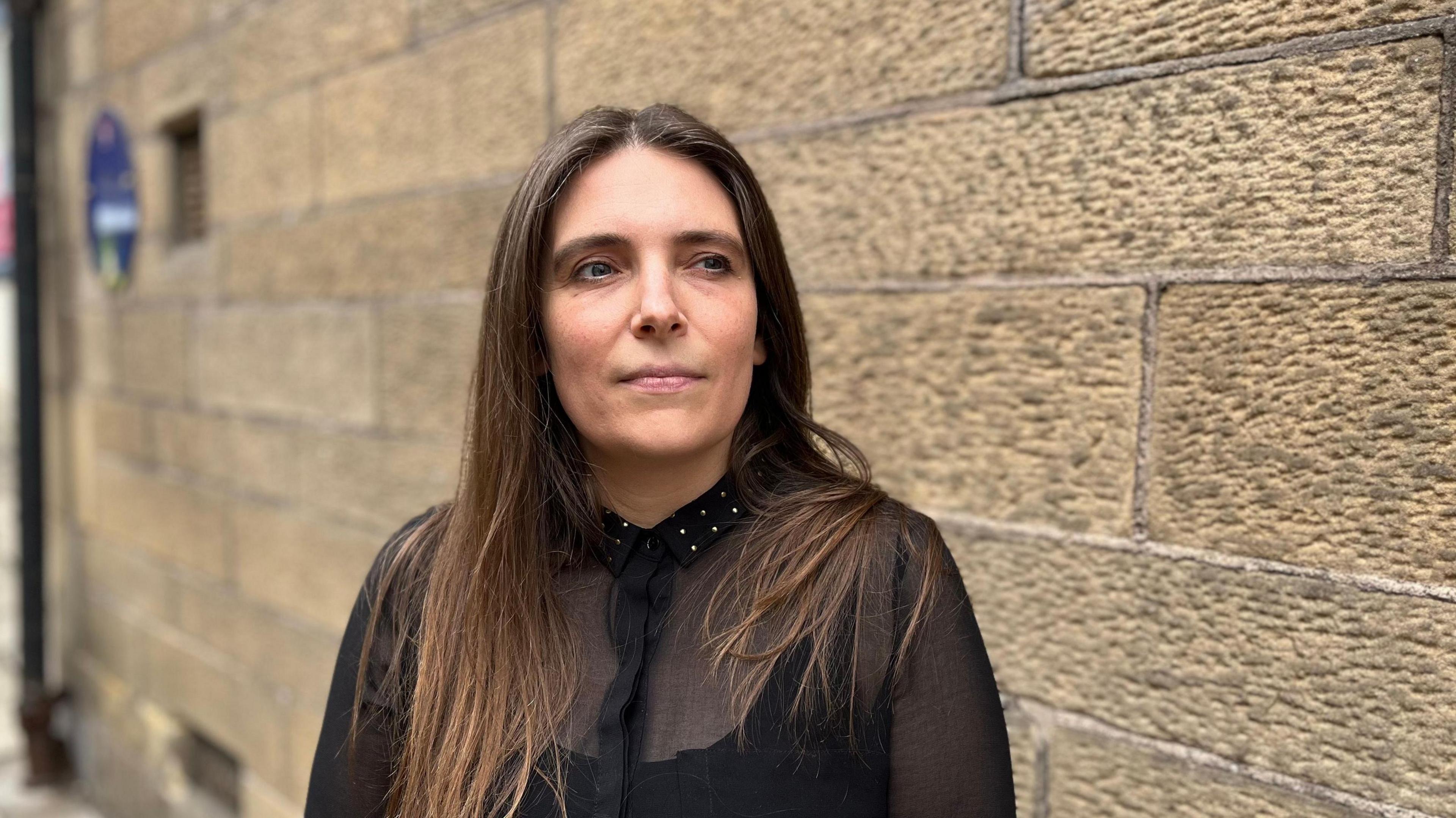 Jenni has long dark hair and a black top. She is standing against a sandstone exterior wall of the building in Jedburgh.
