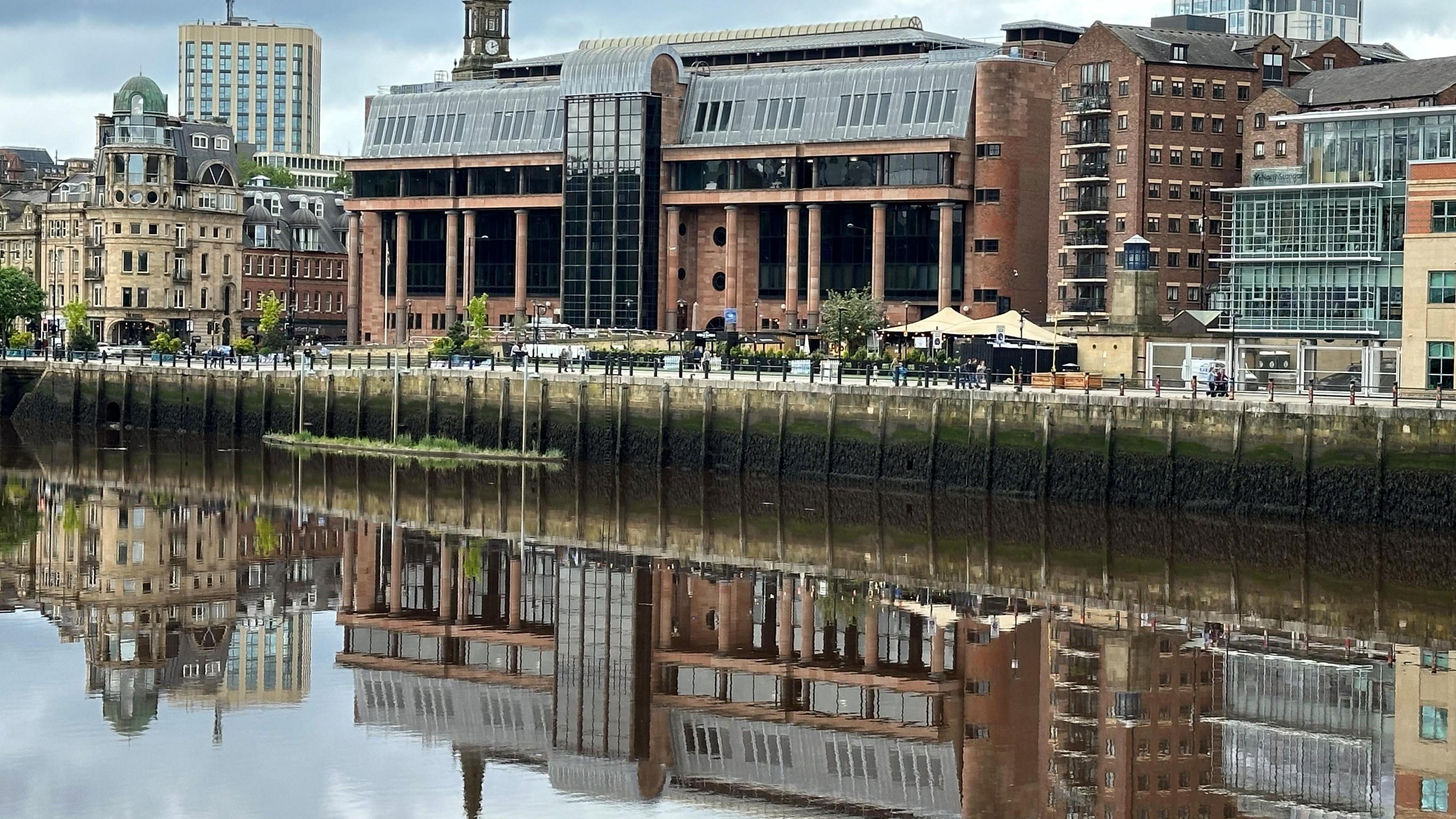 Newcastle Crown Court reflected in the river running in front of it. It is an imposing building of red bricks and columns with large dark windows.