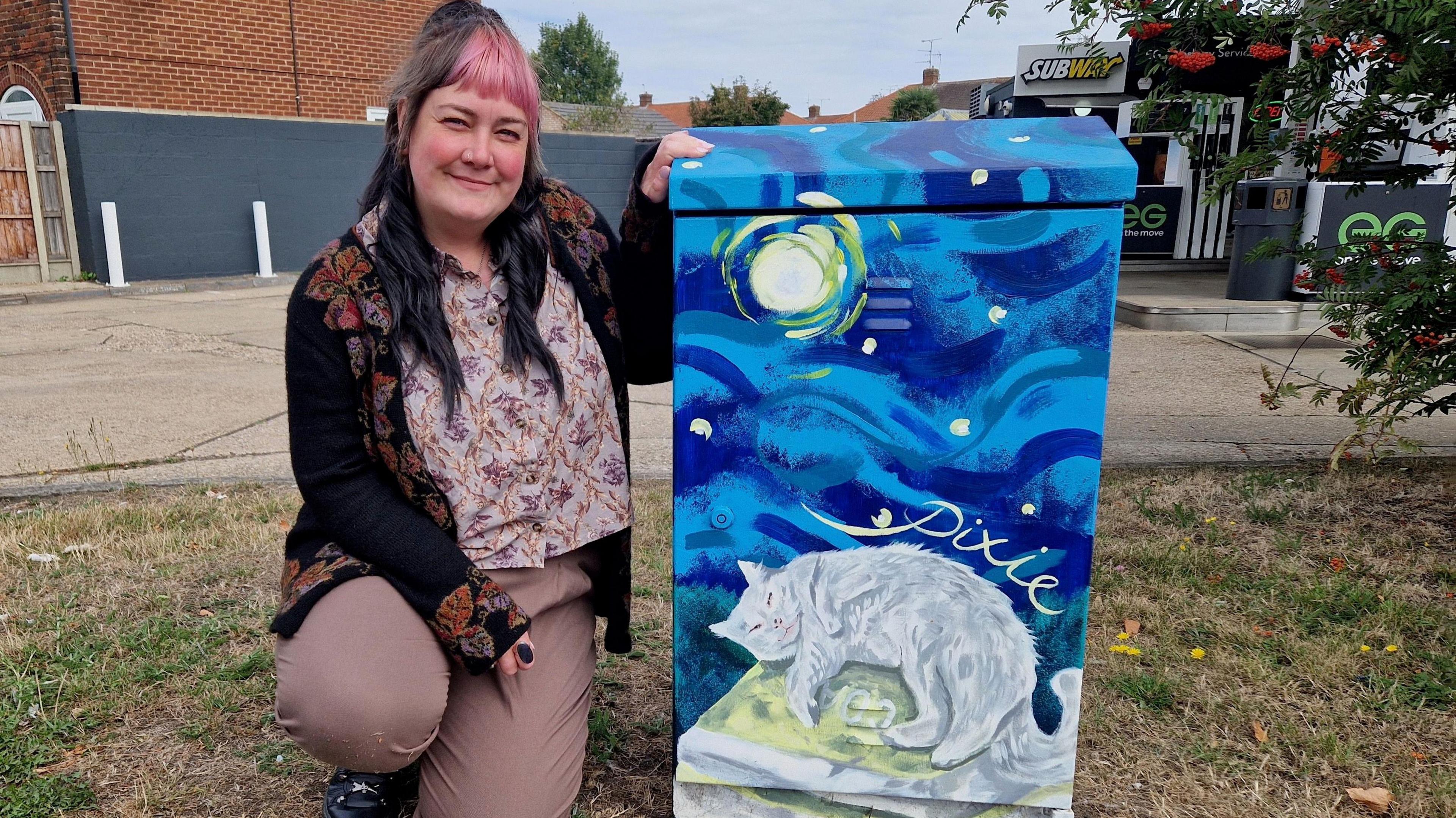 Jo Beck, sitting next to a painted electrical box, showing a blue sky, with a moon and a white cat, sitting on a bag of coals. Her name is painted above her image. There is grass around her and the box and a petrol station in the distance. Jo has dark long hair and a pink fringe, she is wearing pink trousers, a black cardigan and flowery top. She is smiling and looking at the camera.
