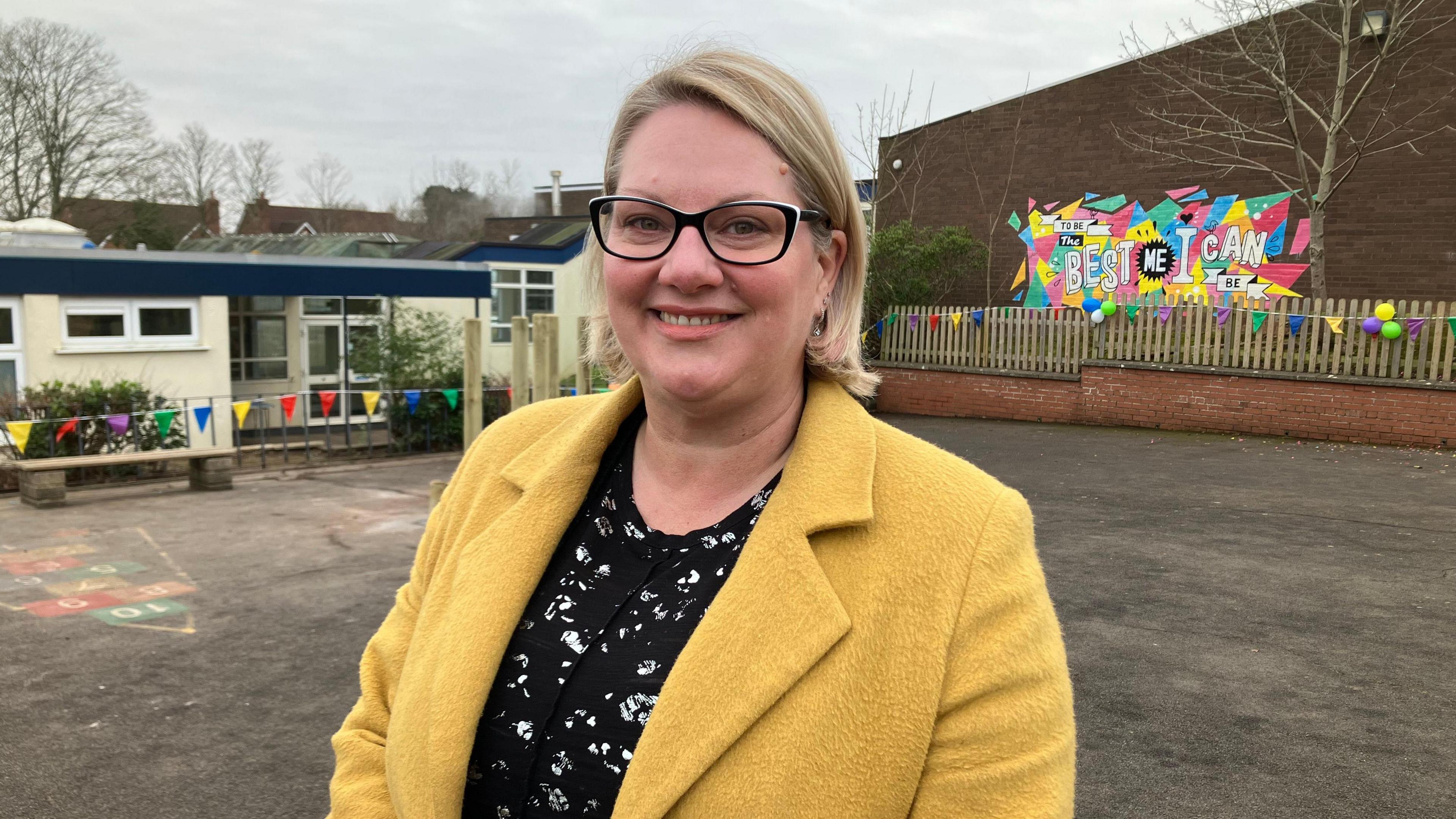 Emma smiles into the camera as she stands in the school playground. She is wearing a yellow jacket, glasses and a black top. Behind her the primary school is visible.