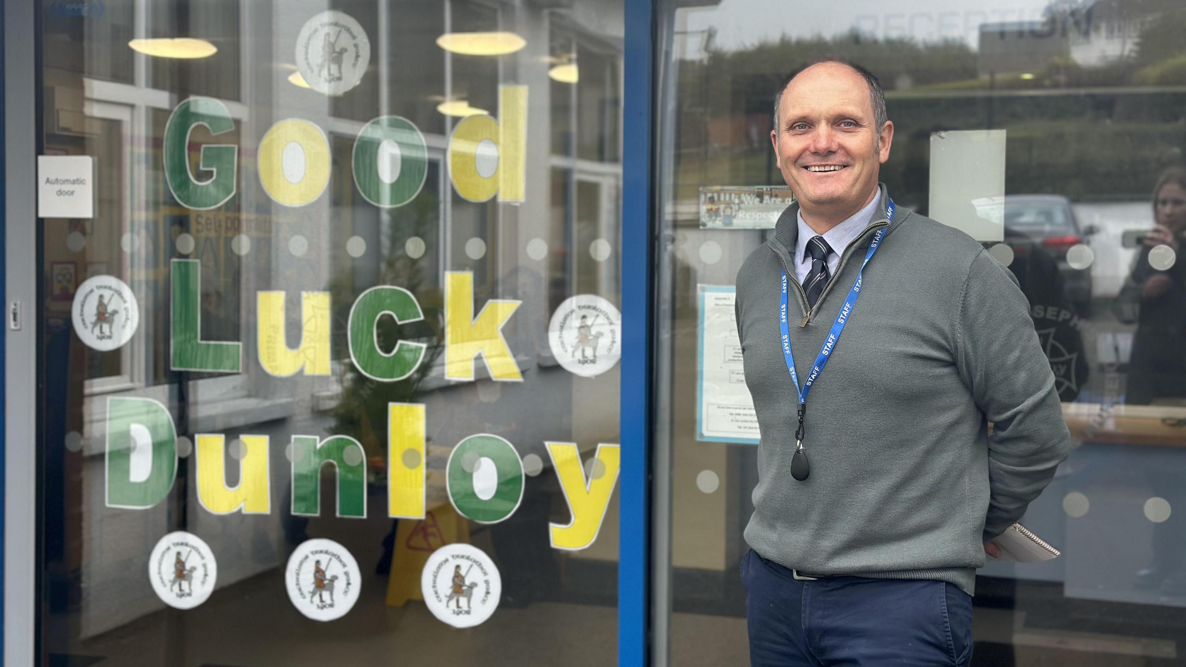A man, wearing navy trousers, a light blue shirt, dark grey tie and a grey jumper, smiling at the camera. He is standing in front of a large glass window that has a sign reading 'good luck Dunloy' on it in green and yellow writing.