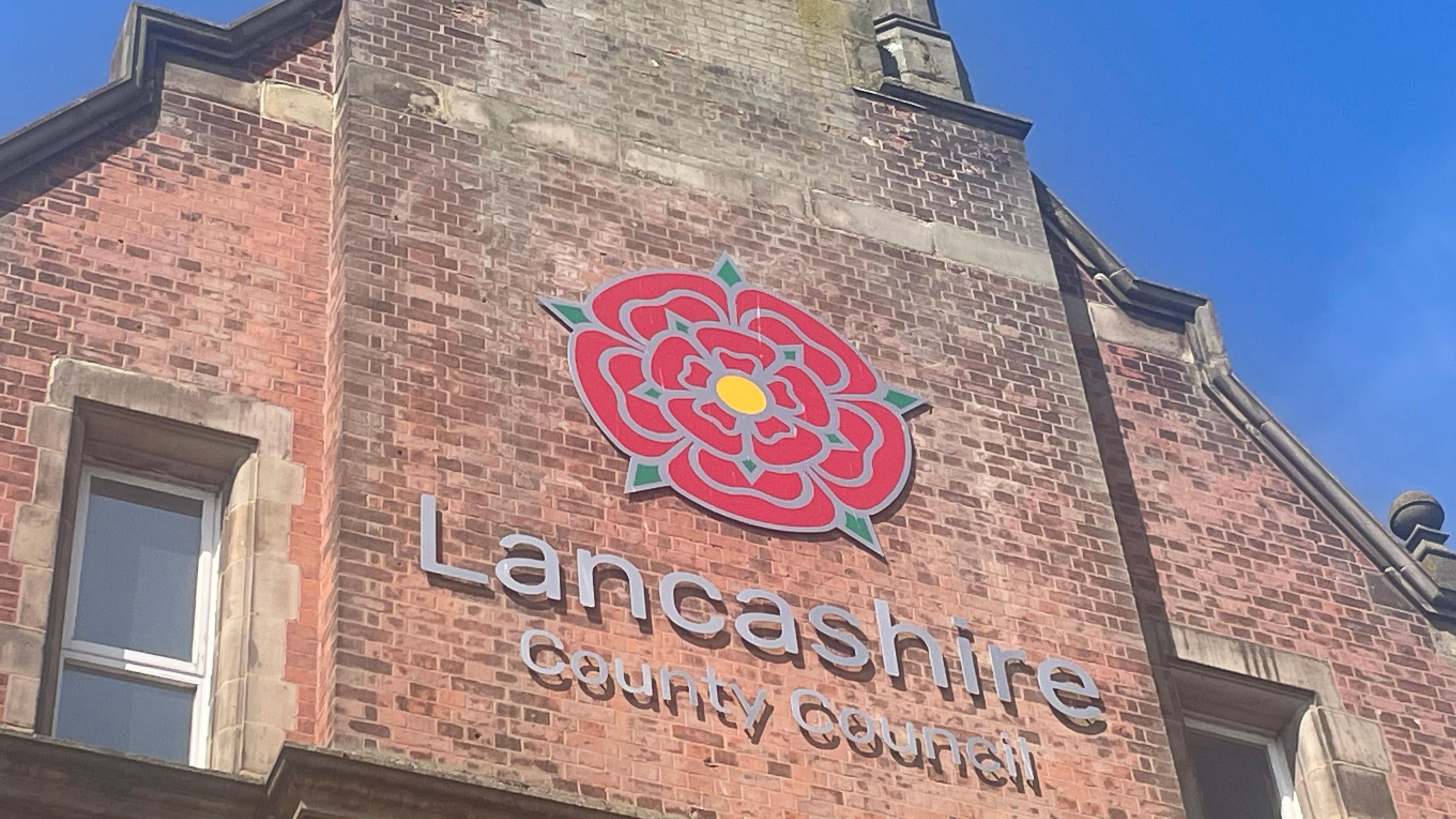 Lancashire County Council sign, with the red rose emblem, on the side of a tall, redbrick building. The photograph was taken on a sunny day with clear blue skies.