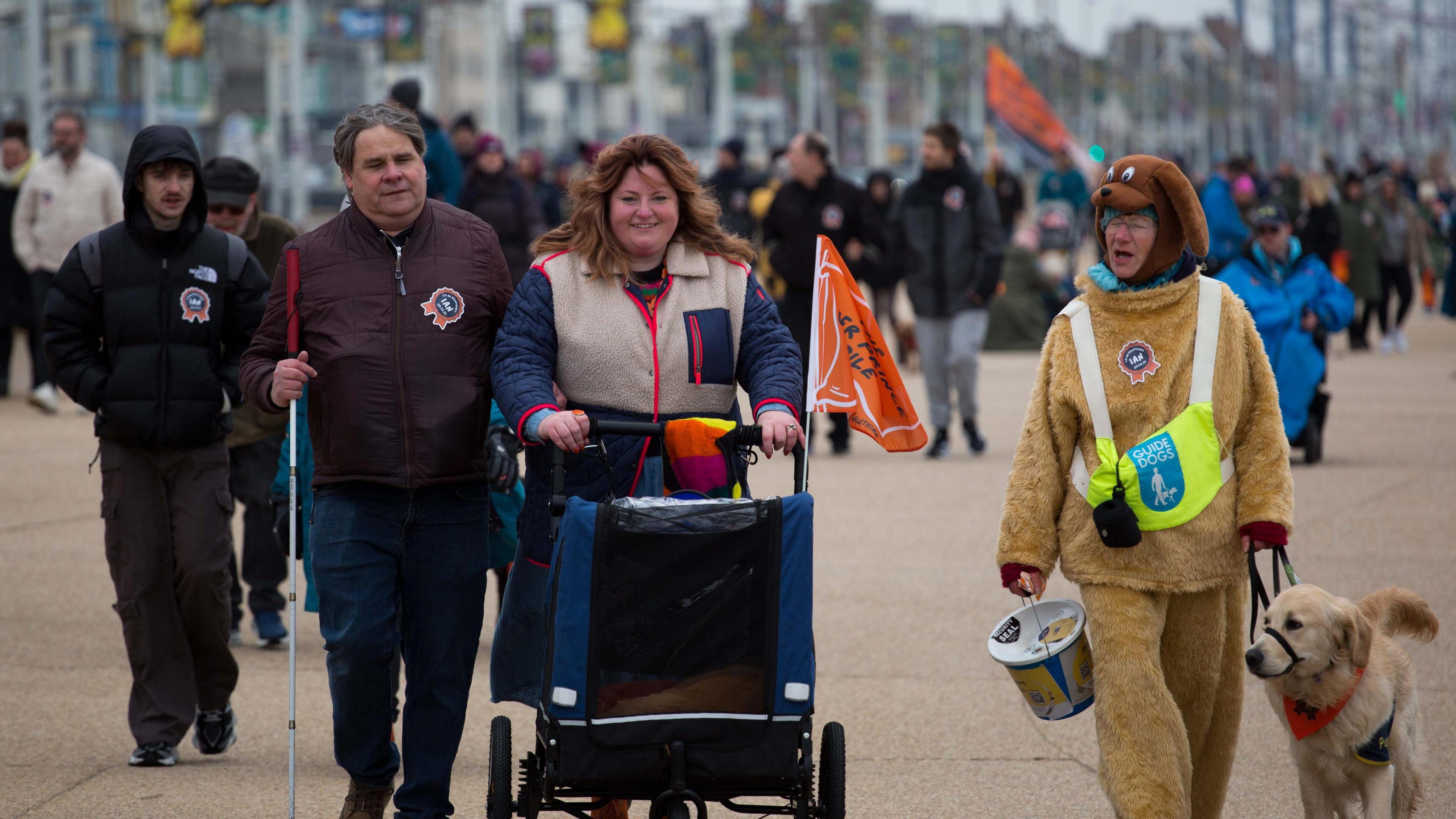 Guide dog user Mark Fielding, with guide dog trainer Gemma Fairhurst, push Ian in a pram along Blackpool Promenade. A charity fundraiser, dressed in a dog costume, holds a charity collection bucket as she walks alongside them.