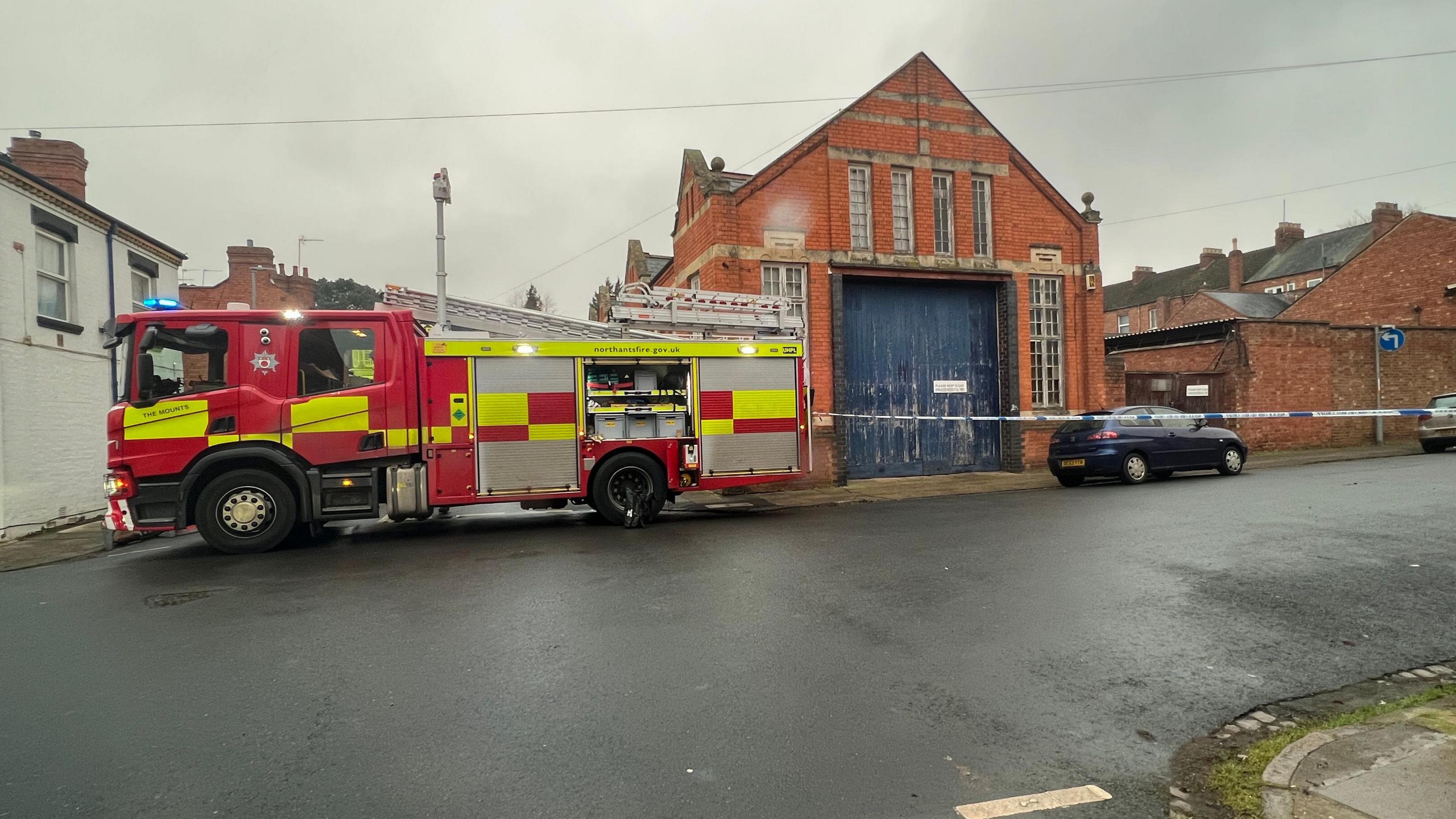 A fire engine parked next to an old industrial-type building with large blue wooden doors. There is a cordon going across the street.