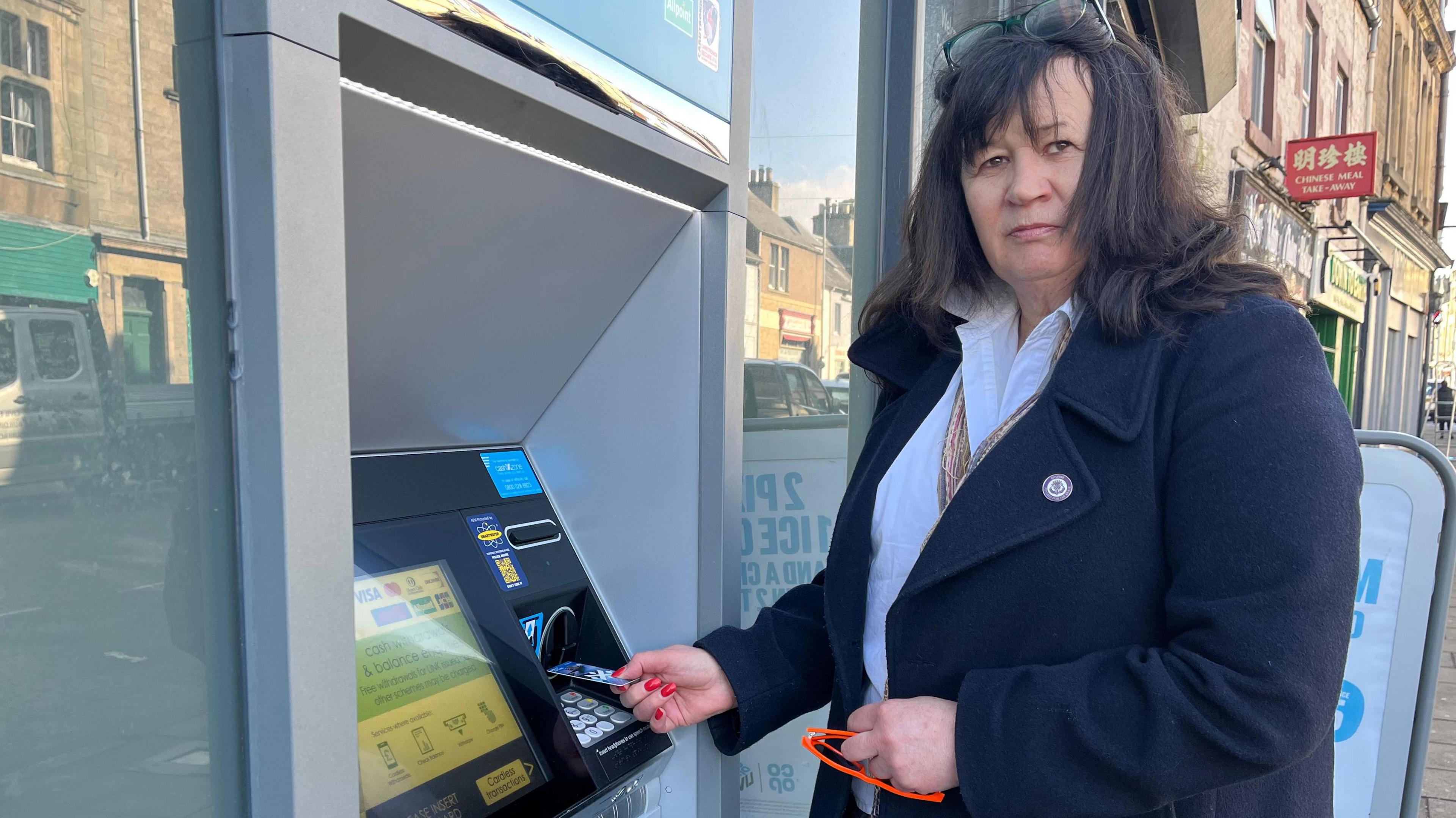 A dark-haired woman with white blouse and black jacket placing a bank card into  an ATM machine