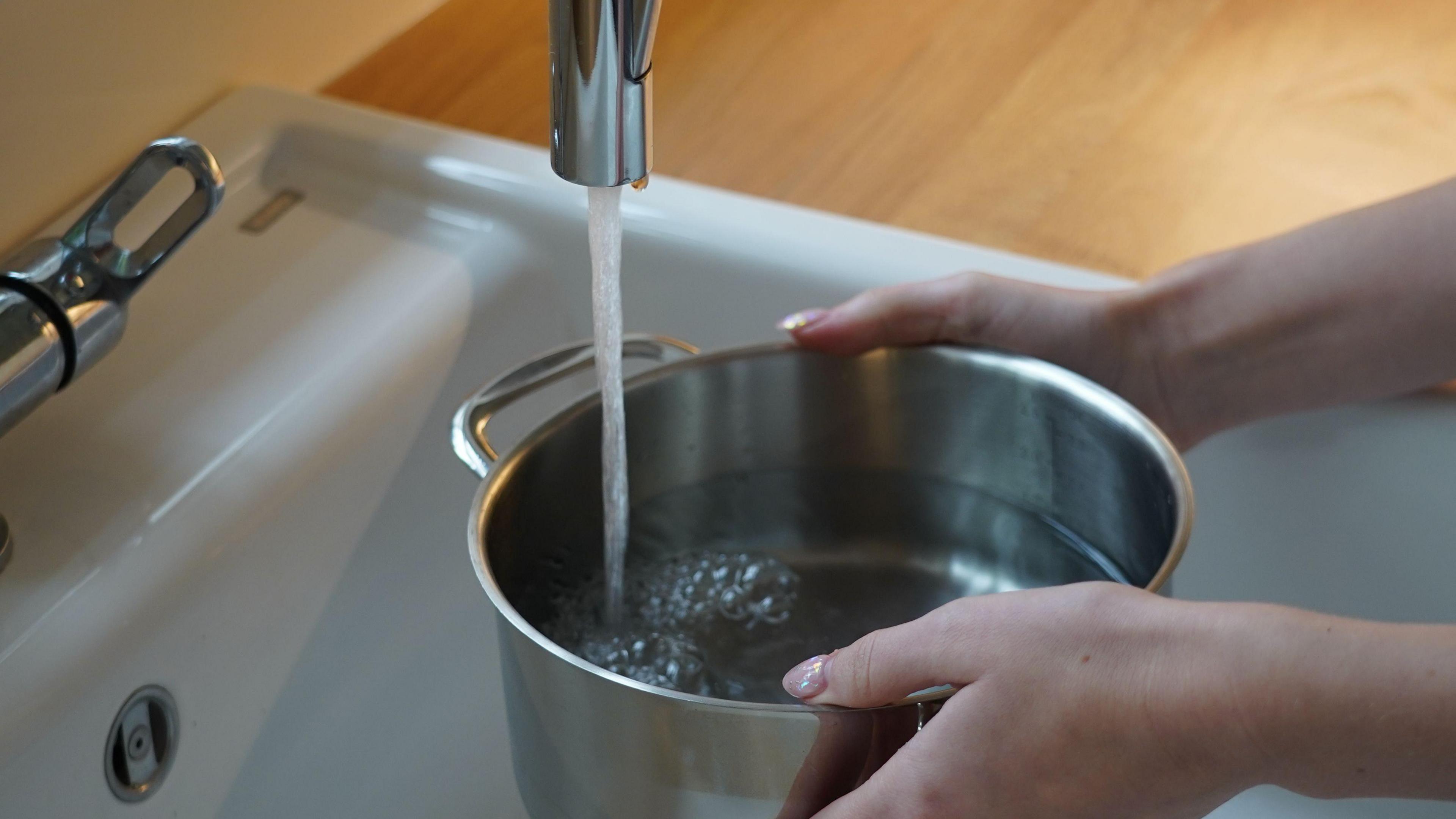 A person filling a pan from a water tap. Only the person's hands are visible and are holding either side of the pan. It is silver and about half full. A stream of water is coming out a silver tap above and the pan is being held over a white sink.