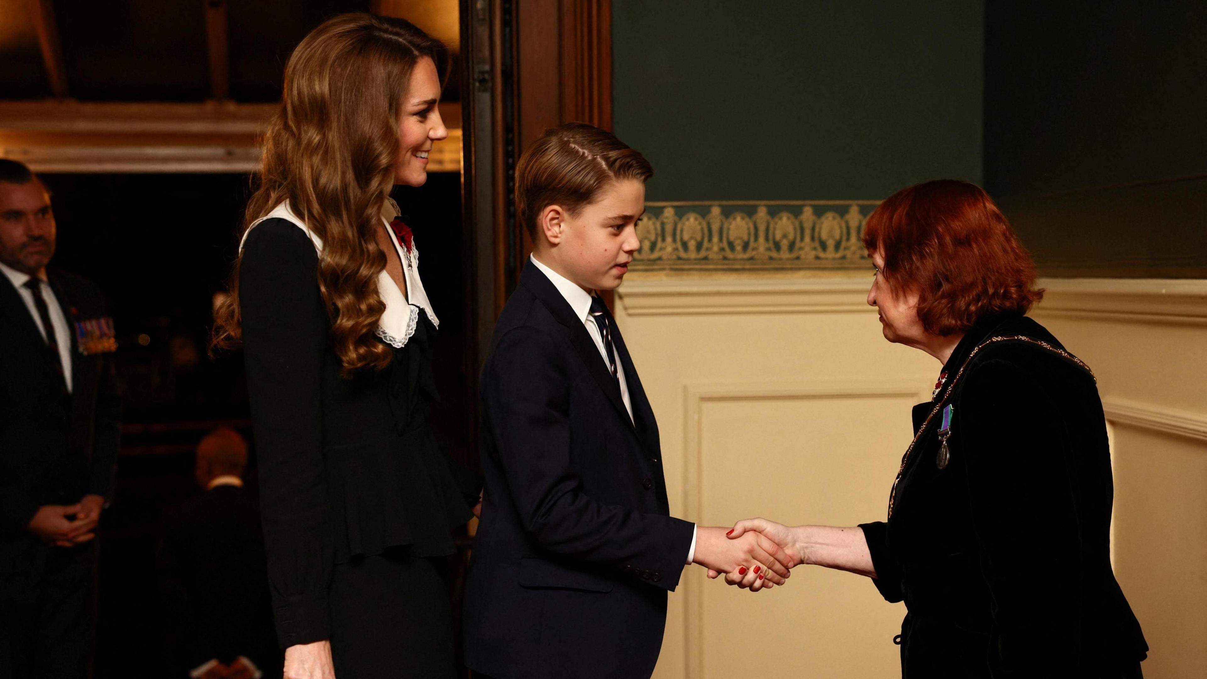 George and Catherine greet a short woman with red hair as they arrive at the concert hall.