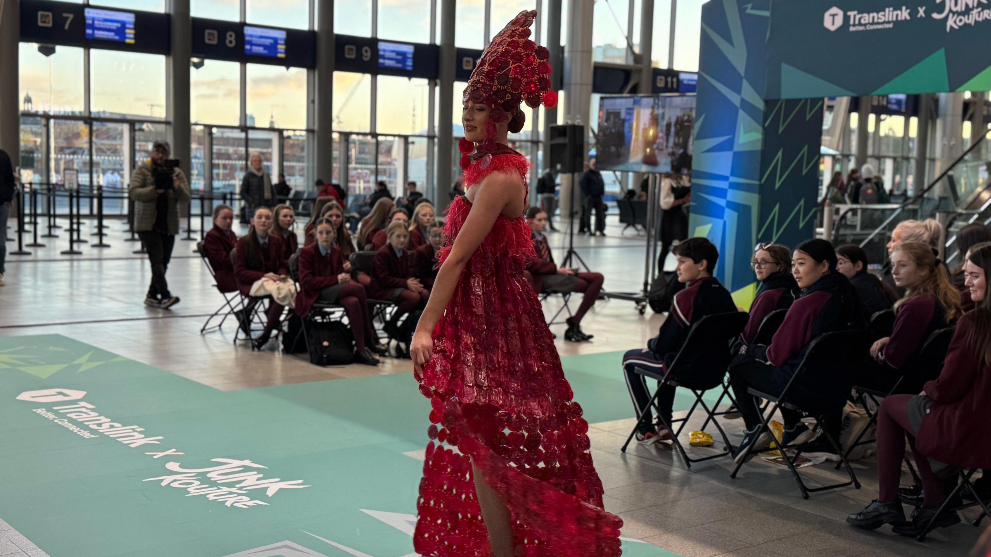 A girl wearing a red dress made of plastic bottles and caps. She is walking the runway. 
