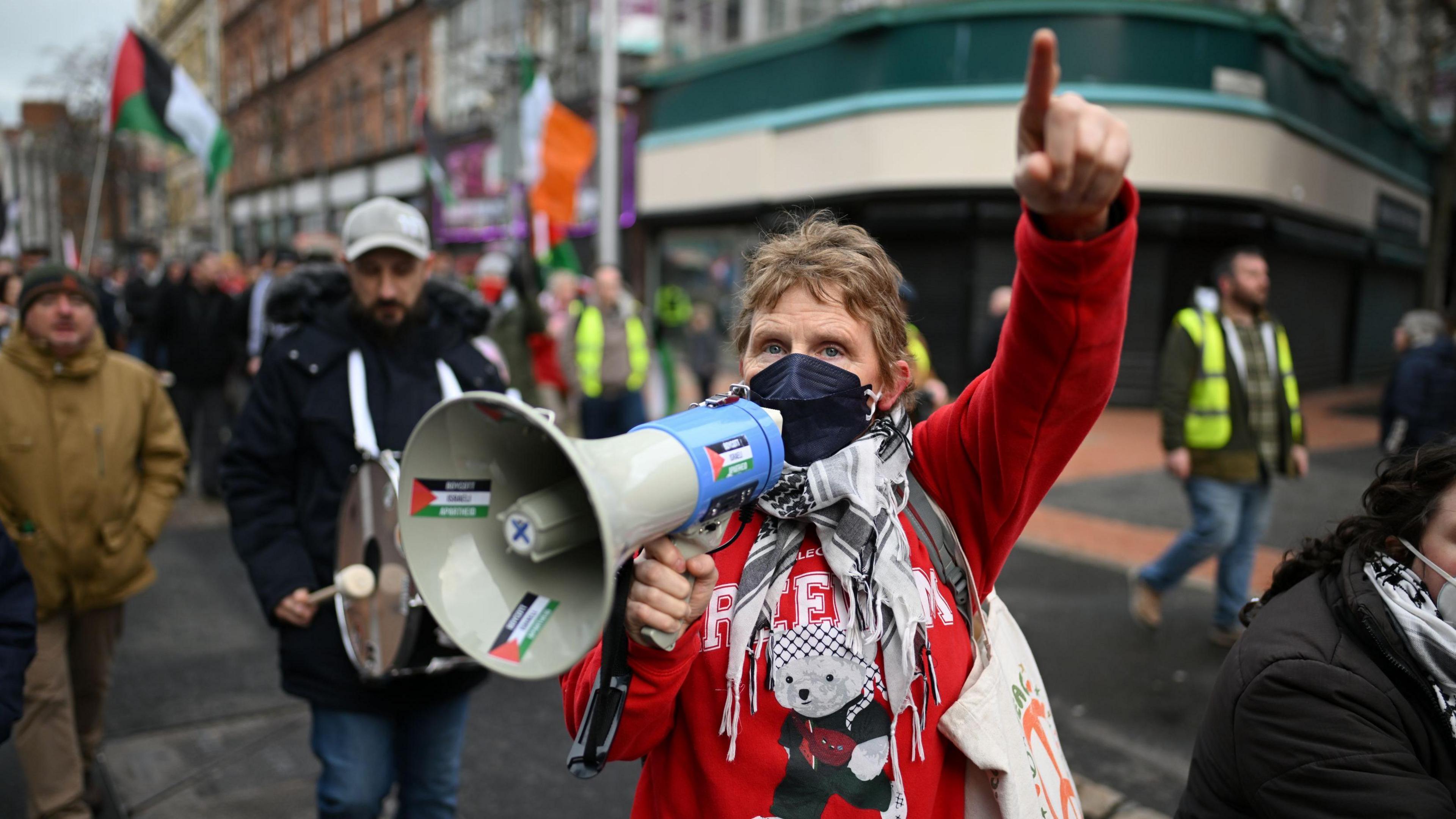 An older woman with short dirty blonde hair in a red top wearing a face mask and scarf is speaking into a megaphone and pointing. The megaphone has Palestine flags on it. Behind her people are walking up in the street in a procession. It is winter in Belfast, many have coats on and a Palestine flag and Irish tricolor can be seen. 