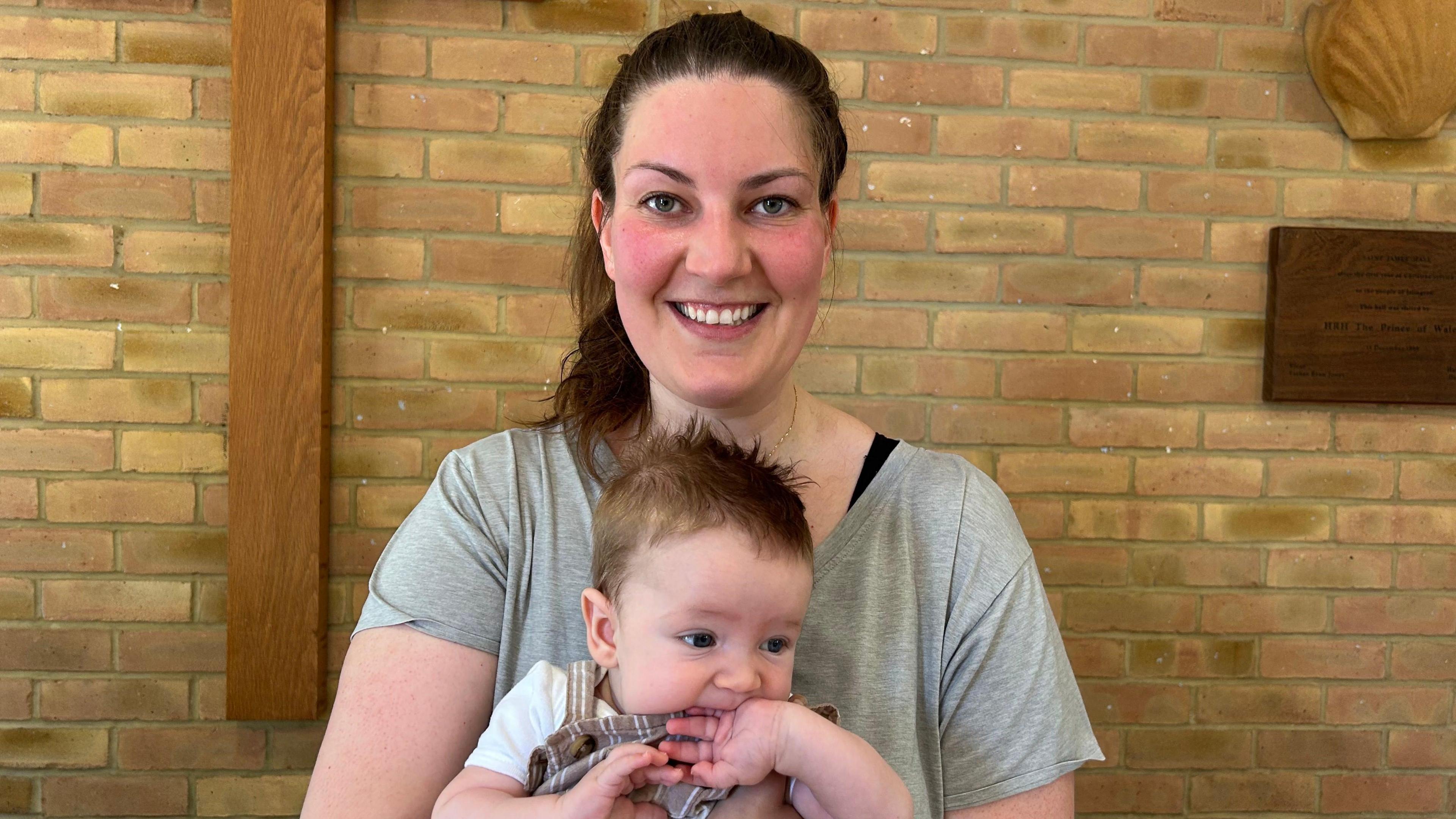 Woman with dark hair tied back in a pony tail, wearing a grey t-shirt and holding a baby on her lap with dark brown hair and wearing a pinafore top and white t-shirt. Woman smiles at the camera.