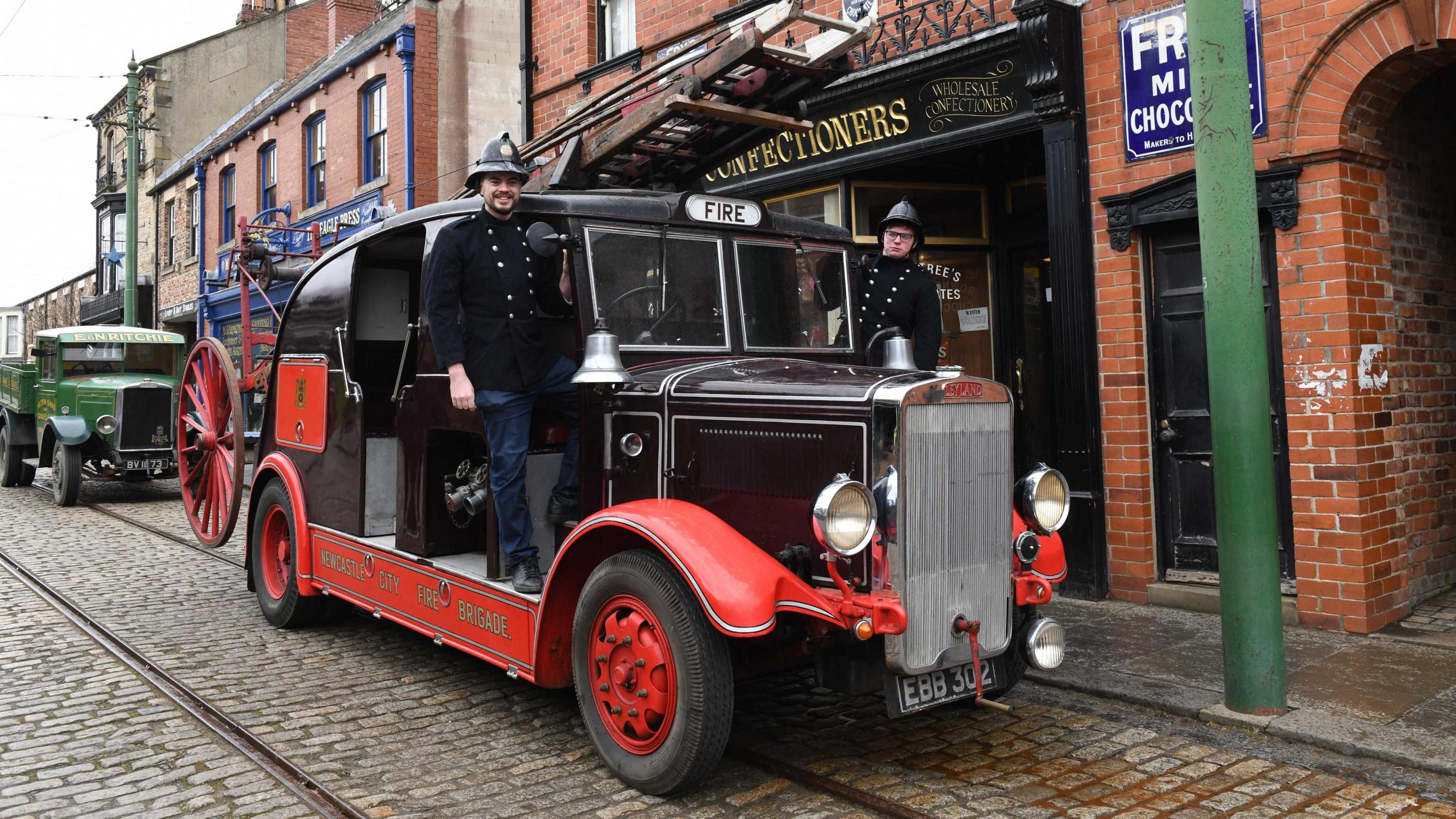 Jonny and Sam from Beamish Museum of the North pictured on the fire engine after it arrived at the museum. The vehicle is black and red and parked on a cobblestone road which has tram tracks running along it. The people on the truck are wearing old-fashioned black helmets and uniforms. 