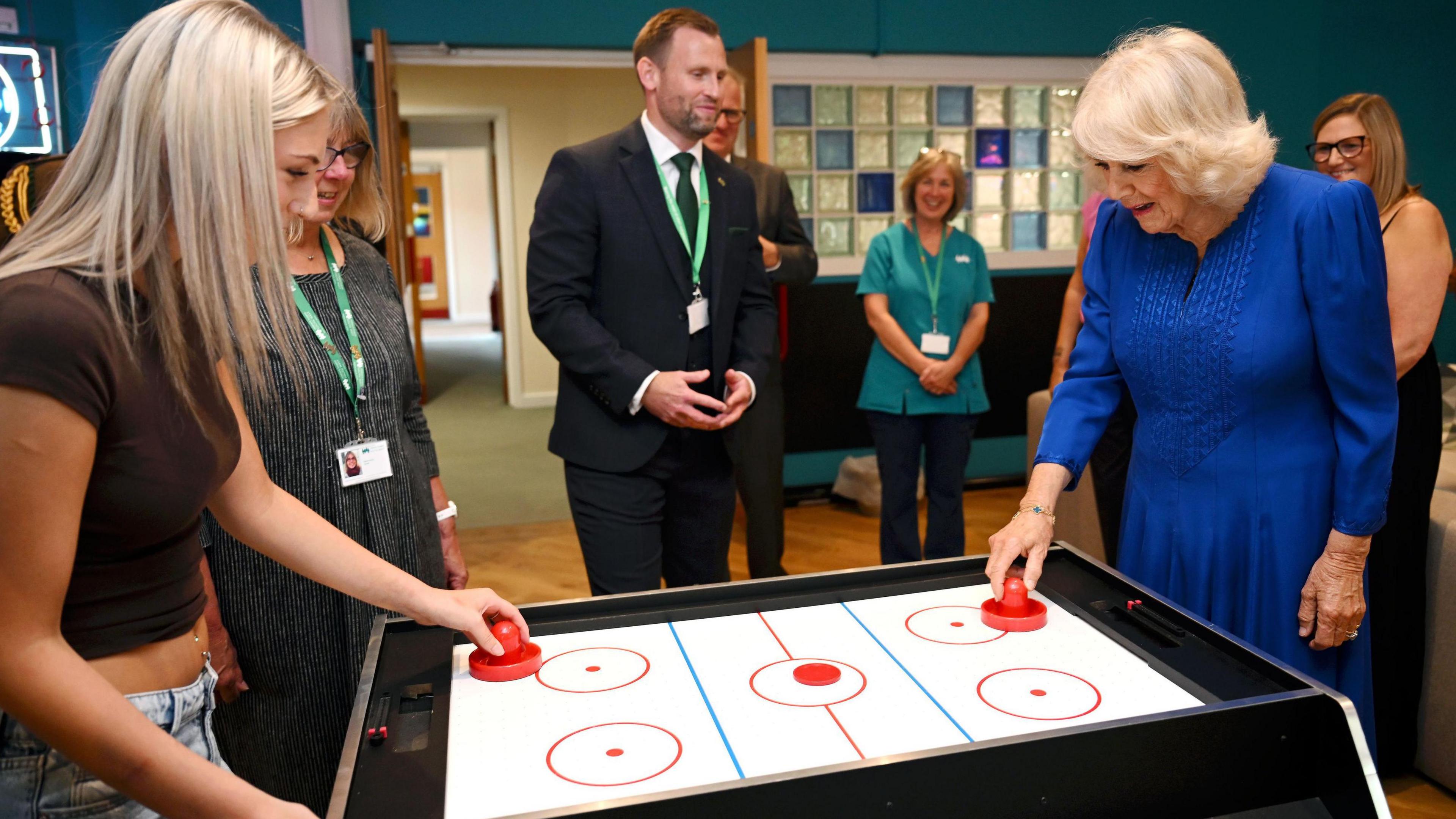 Queen Camilla, a woman with shoulder length grey hair, wearing a blue dress, plays air hockey with a teenage girl, with bleached blonde hair, wearing a brown crop top and jeans.