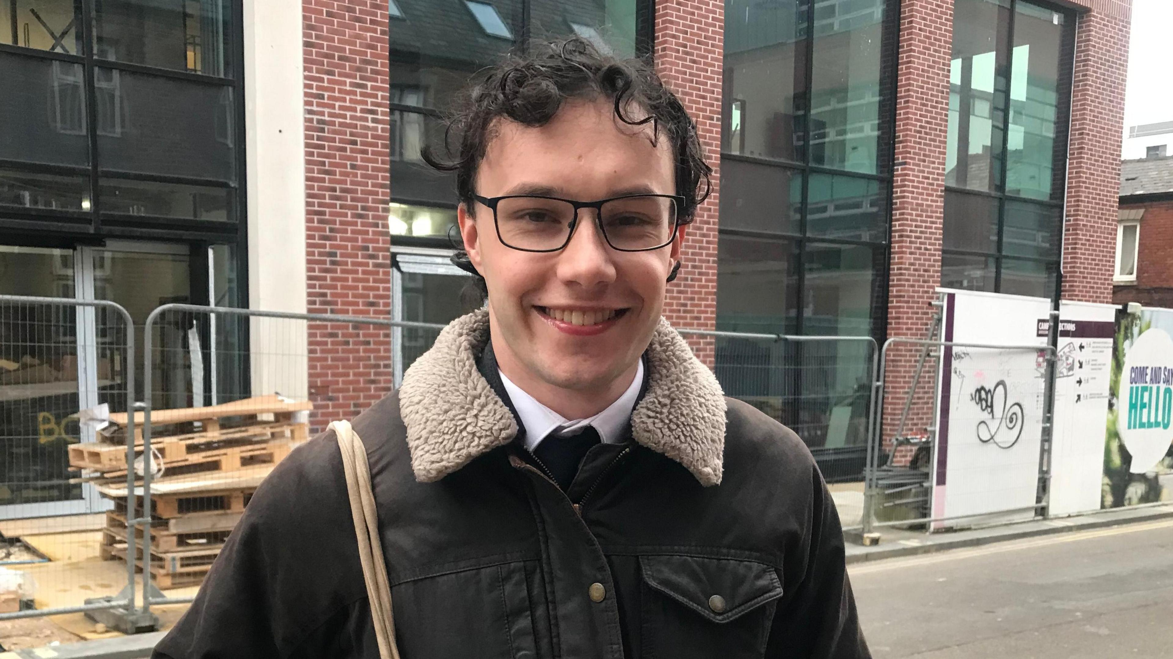 Mr Richardson stands in front of a construction site, wearing a jacket and glasses. He has a tote bag slung over his right shoulder and smiles at the camera.