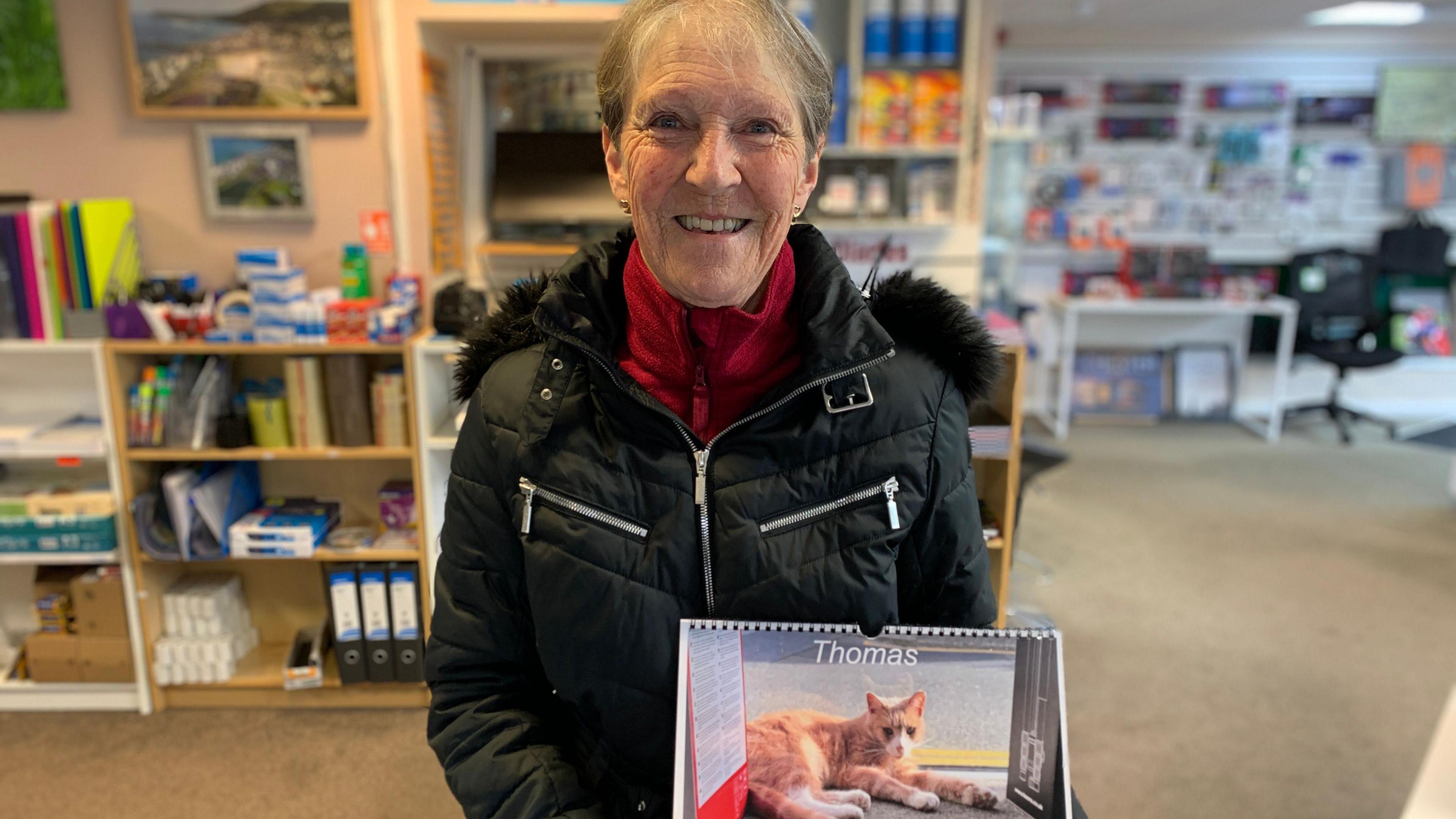 A woman with grey-brown hair wearing a dark down jacket over a red fleece. She is standing in a stationary shop and holding a wall calendar with an image of the cat on the front, and the title Thomas.