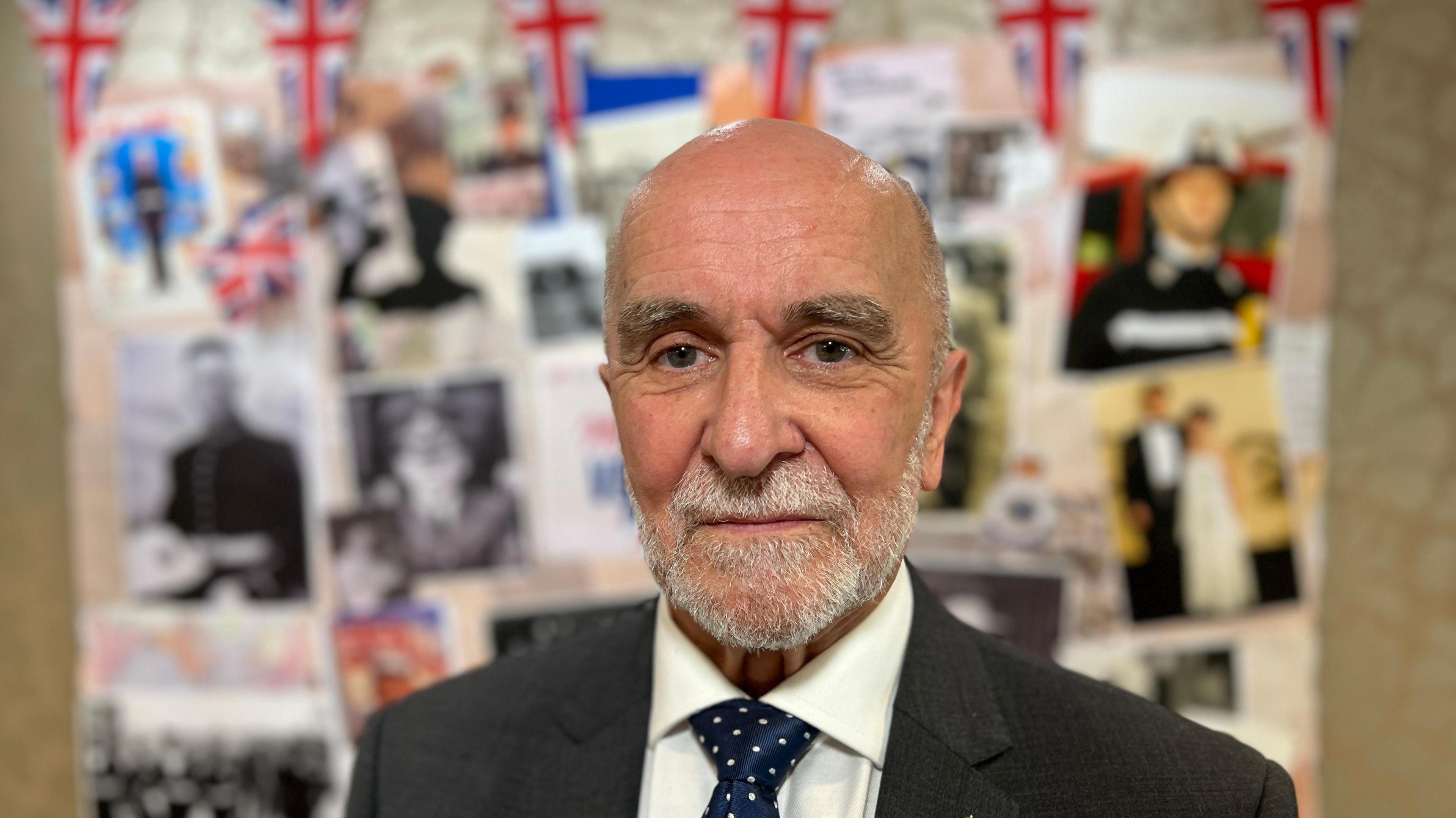 A man in a suit and tie stands before a wall adorned with photographs and Union Jack flags. The backdrop features images of individuals in military dress, formal wear, and other commemorative or historical scenes