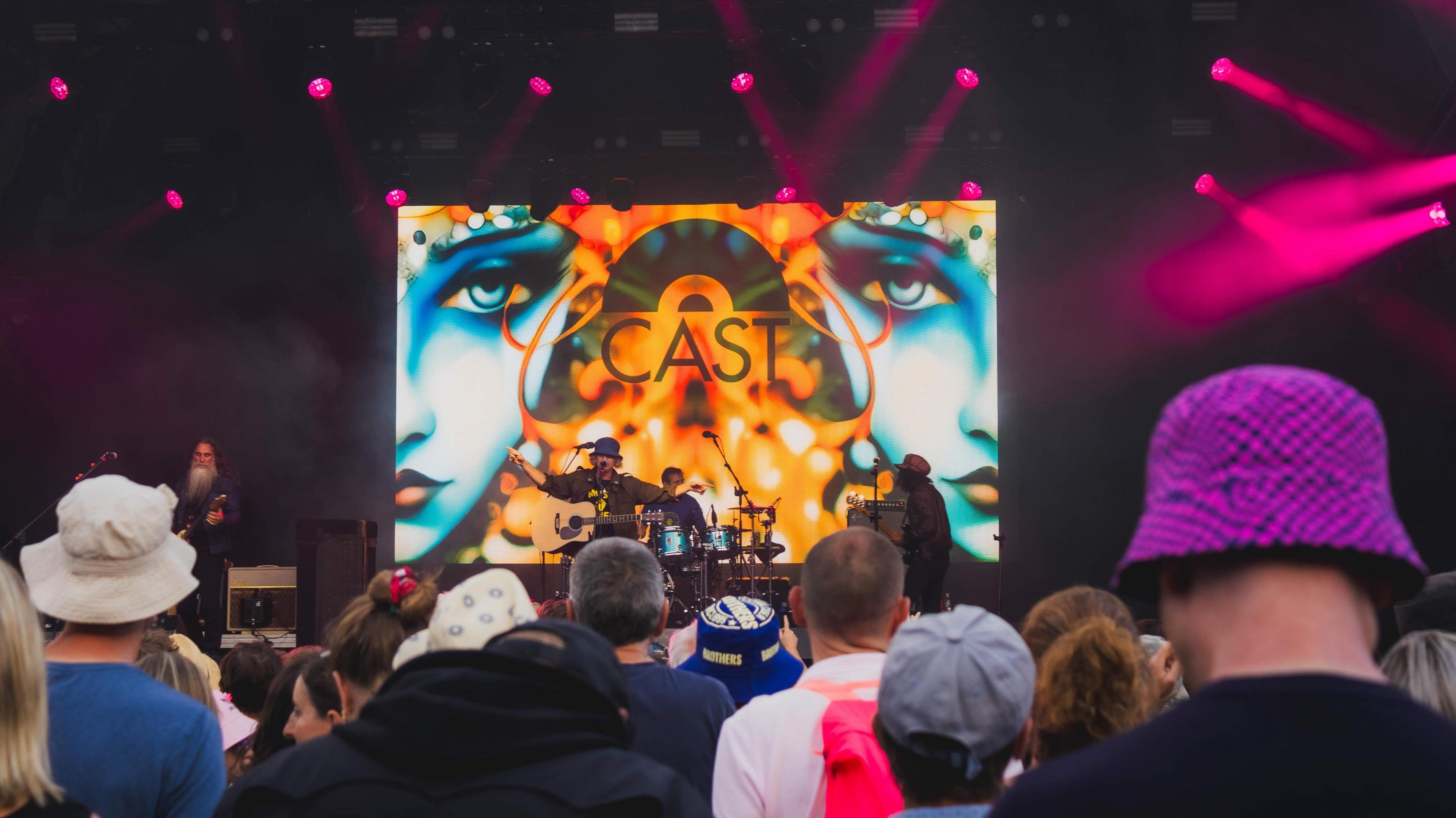 Cast performing at Bristol Sounds. The picture is taken from the crowd. Backs of people's heads can be seen and the band is in the distance. The stage is orange with two blue faces projected on the back.