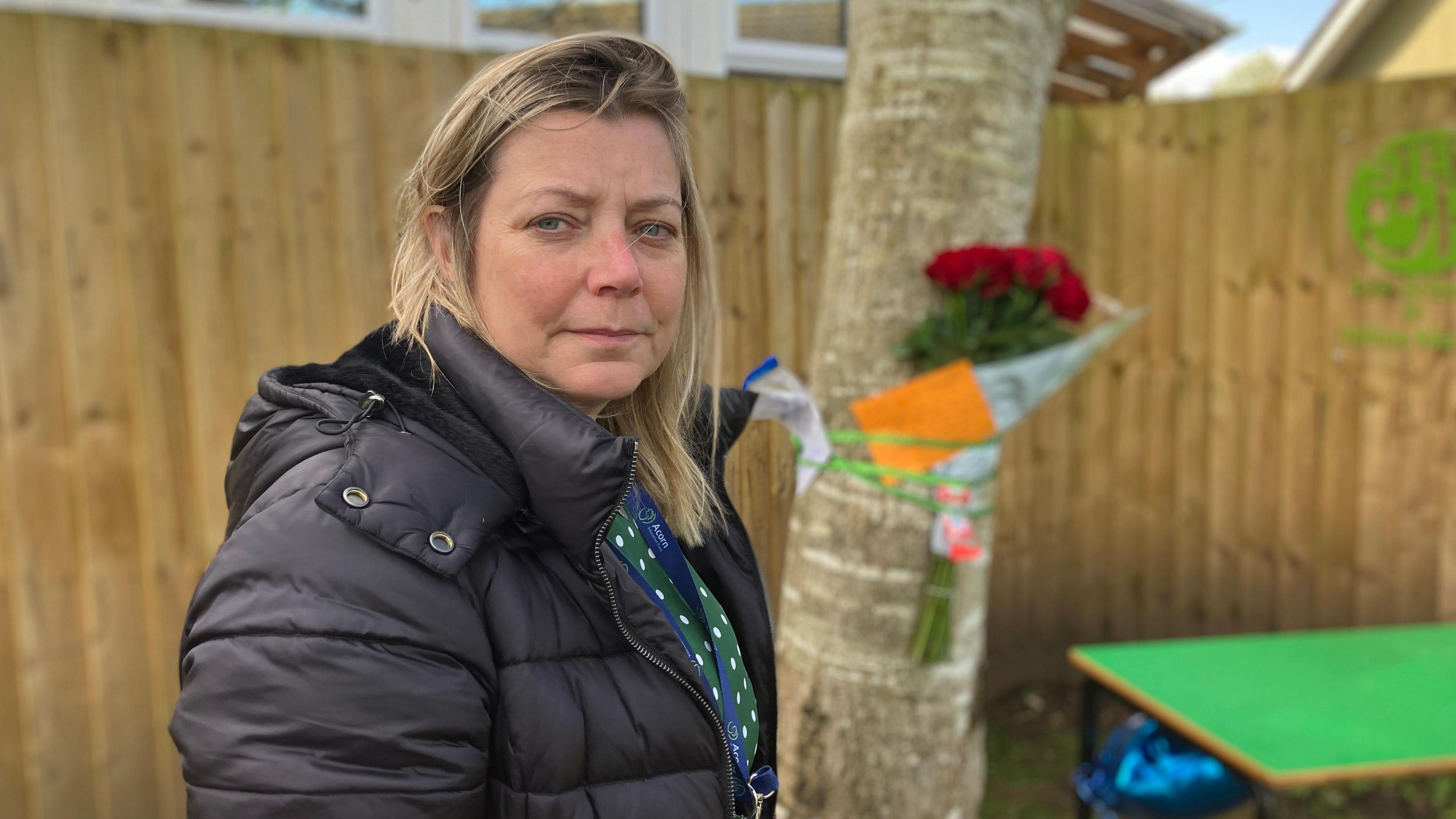 A picture shows the headteacher of Dilton Marsh Primary School, Jill Hibbs, standing outside in a black coat in front of a tree. The tree has a number of tributes including cards and flowers tied to it with green ribbon. 