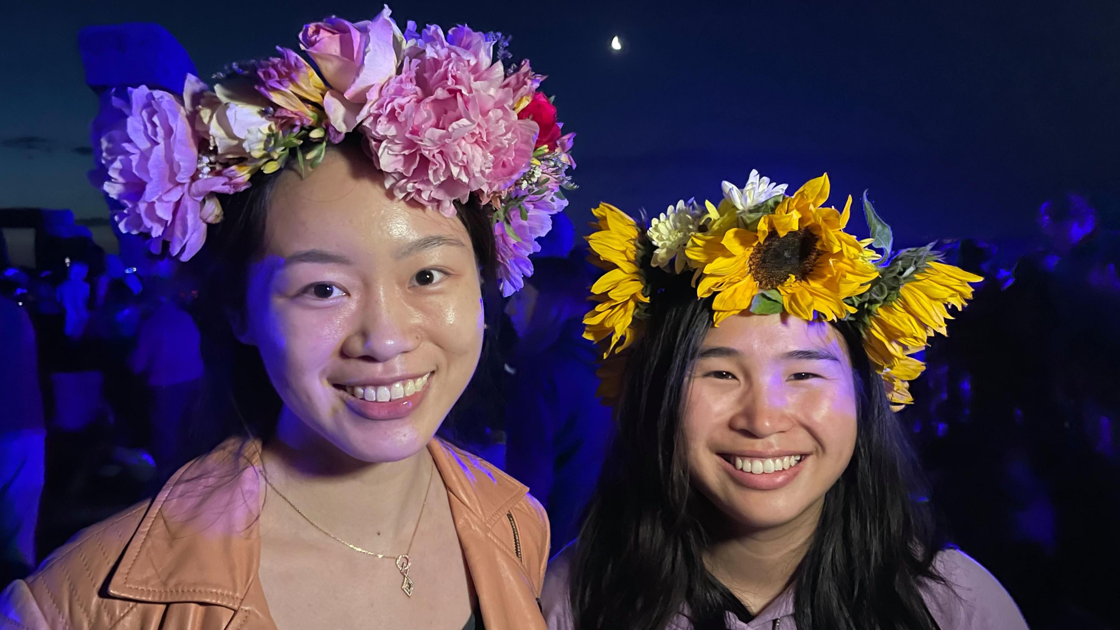 Two women smililng into the the camera. Both have flower headwear. One is purple, the other is yellow. One of the women is wearing a light brown coat, the other is wearing a purple top.