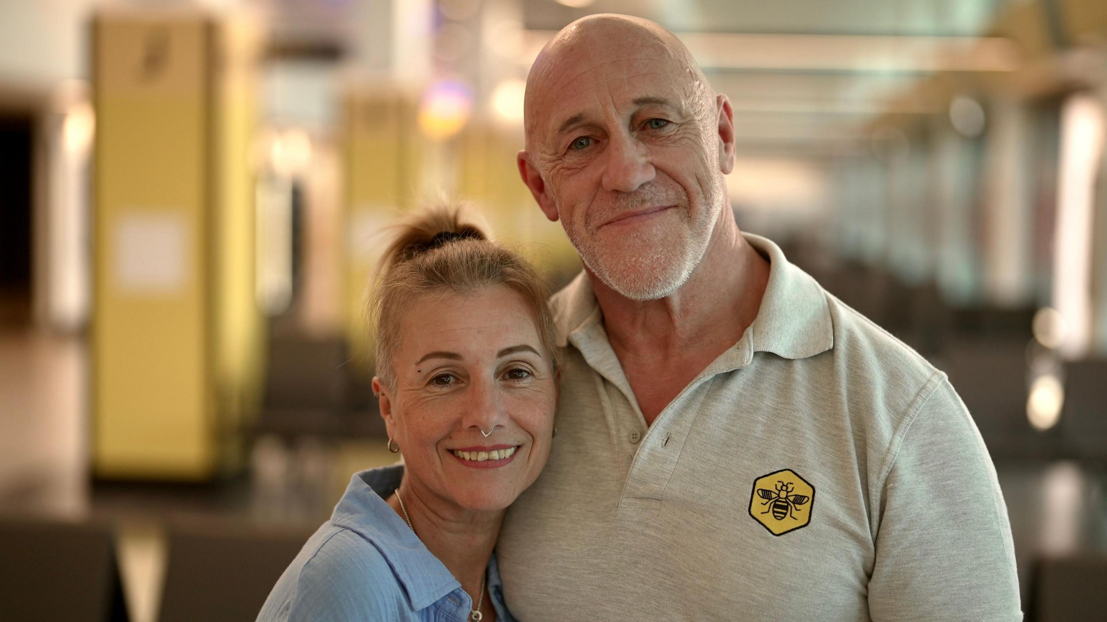 Two adults standing close together in the foreground of an indoor waiting area. One person wears a light blue shirt over a white top and has hair tied back; the other wears a light grey polo shirt with a yellow badge featuring a bee symbol and has a tattooed forearm. Rows of dark chairs are visible behind them, with blurred columns, ceiling lights and windows extending along the corridor in the background. No visible bags, bins or markers are present.