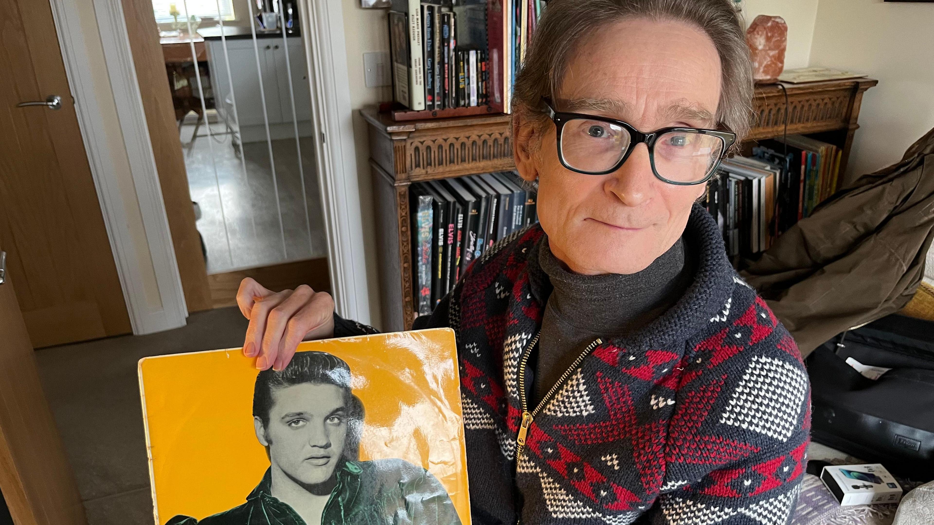 Giles Moriarty, 57 is wearing glasses and sitting in a chair holding an Elvis Presley album showing the young Elvis in black and white on a yellow background. Giles has brown hair and a dark rolled neck sweater with a red black and white patterned cardigan over the top, there is a dark wooden book case behind him filled with Elvis books.