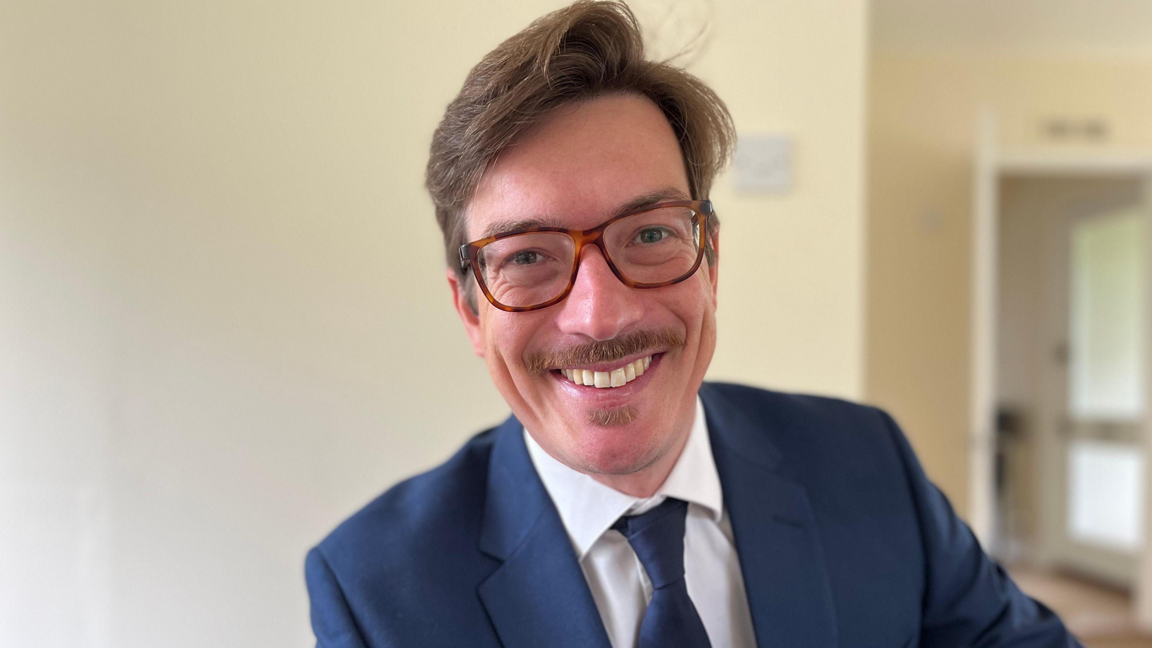 Matthias Plunser, wearing glasses and a moustache, smiling towards the camera. He is wearing a blue suit and tie