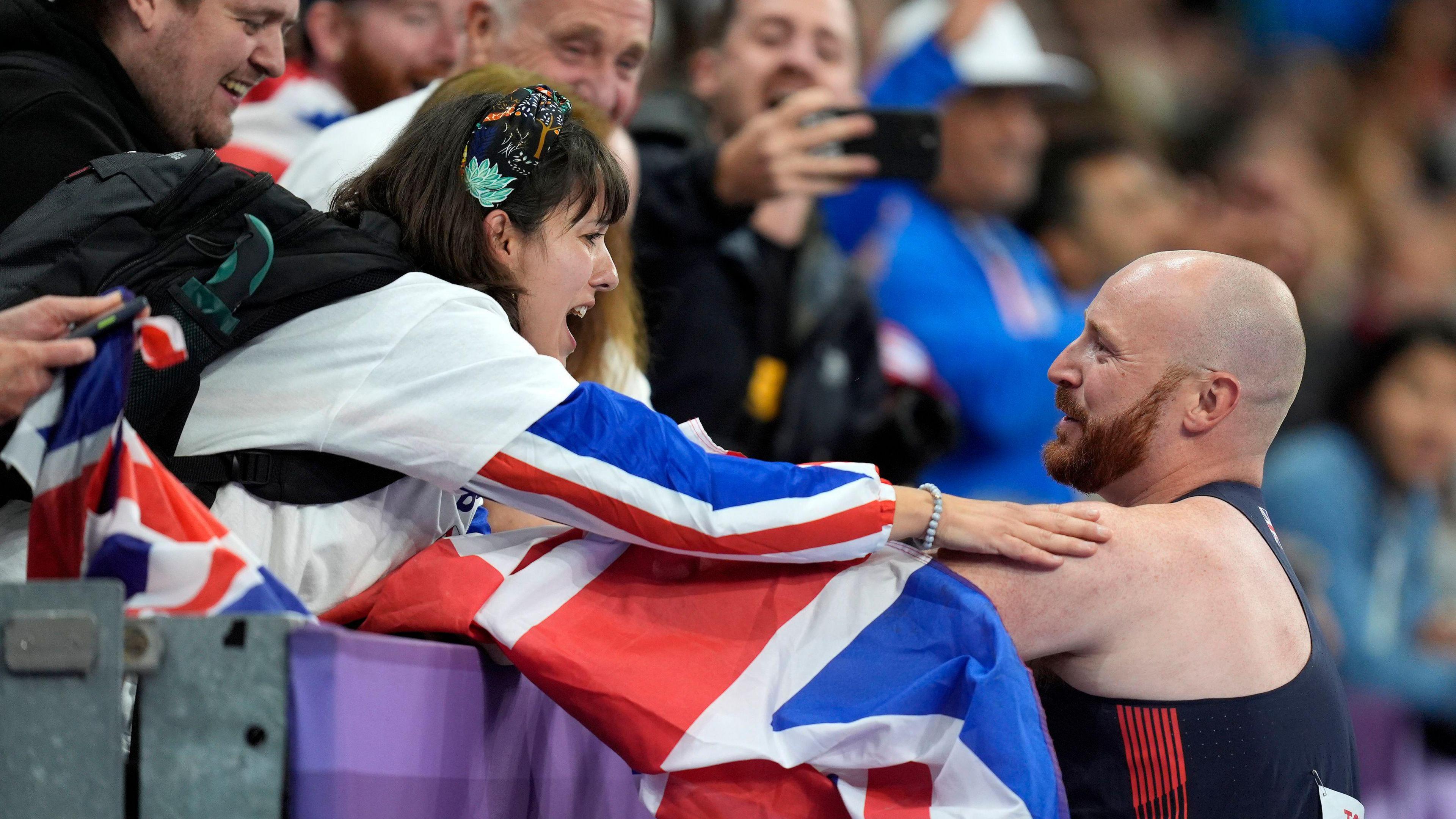 Dan Pembroke reaches out to his wife Martina in the crowd over a union jack draped on the barrier. She has short black hair and a full fringe, resting her arm on his shoulder. The man has a shaved head and brown beard.