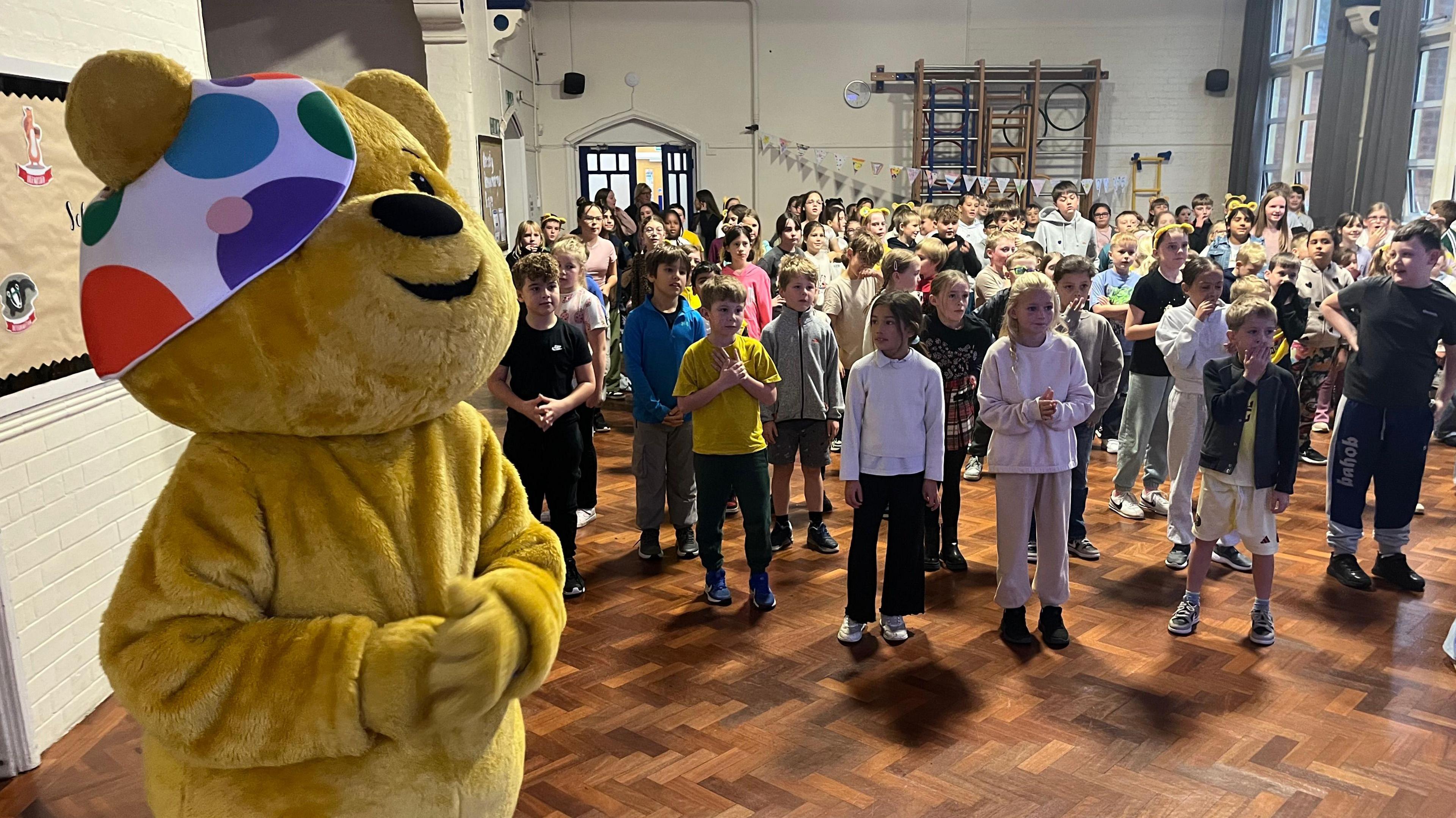 Pudsey Bear stood at the front of a school hall. A large crowd of children are in the distance.