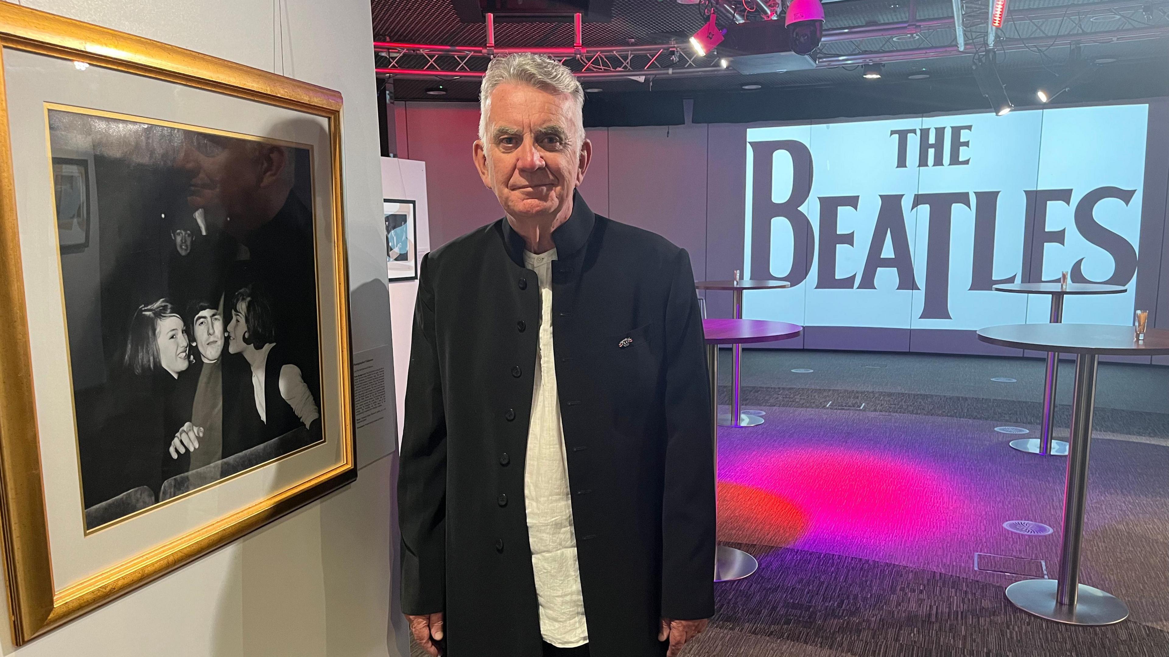 Mark Stanton wearing a black jacket over a white shirt stands in an exhibition space alongside a picture hanging on a white wall, with tables and a large screen in the background behind him with the words 'The Beatles' in large black writing.