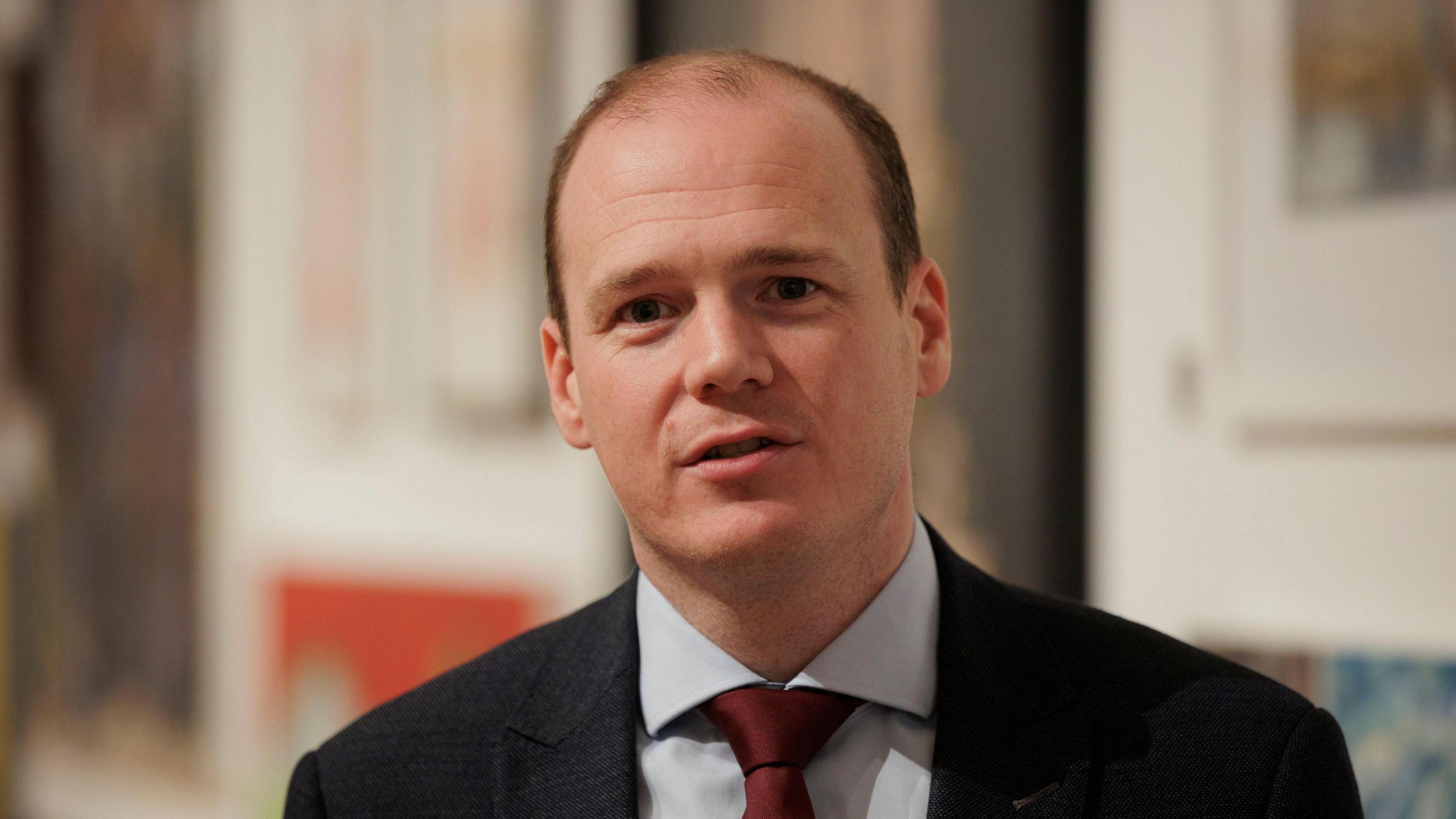 A man with short dark hair is talking to someone off camera. He is wearing a dark-coloured suit jacket, a light shirt and a red tie. The background is out of focus. 