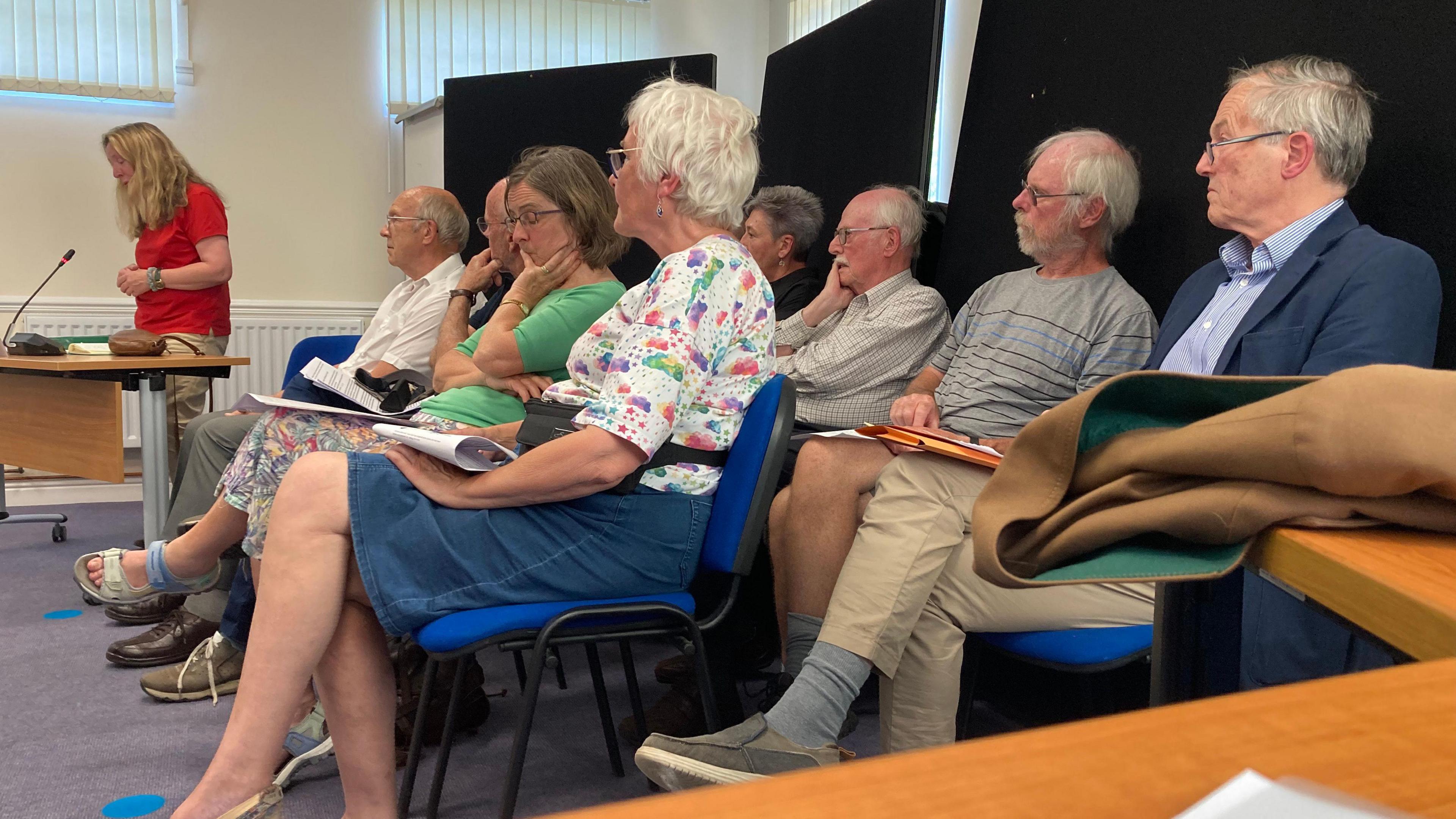 Eight people sit on chairs at a council meeting, listening to a woman who is standing up and talking into a microphone