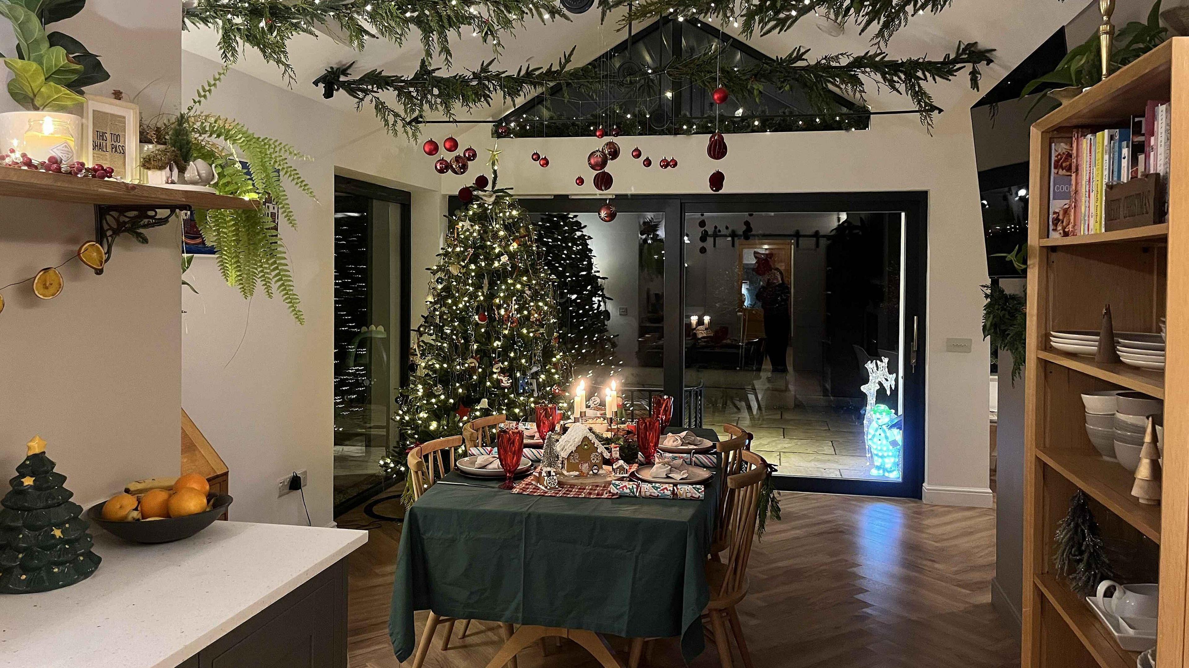 A Christmassy kitchen with a vaulted ceiling. Red Christmas baubles dangle from the ceiling, which is also decorated with Christmas foliage. In the corner is a large Christmas tree decorated with hundreds of Christmas lights. A dining table, with a green tablecloth, is garnished with candles and a gingerbread house. 