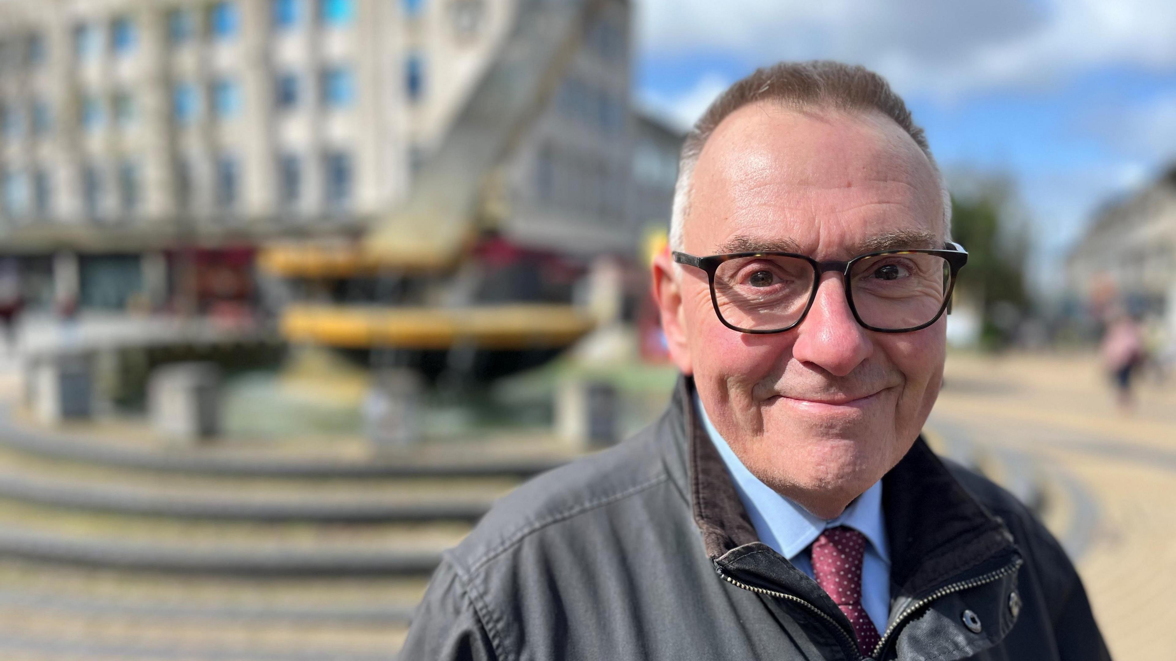 Plymouth City Council leader Tudor Evans wearing a blue shirt, red spotted tie and a brown coat in Plymouth city centre with a blurred background.